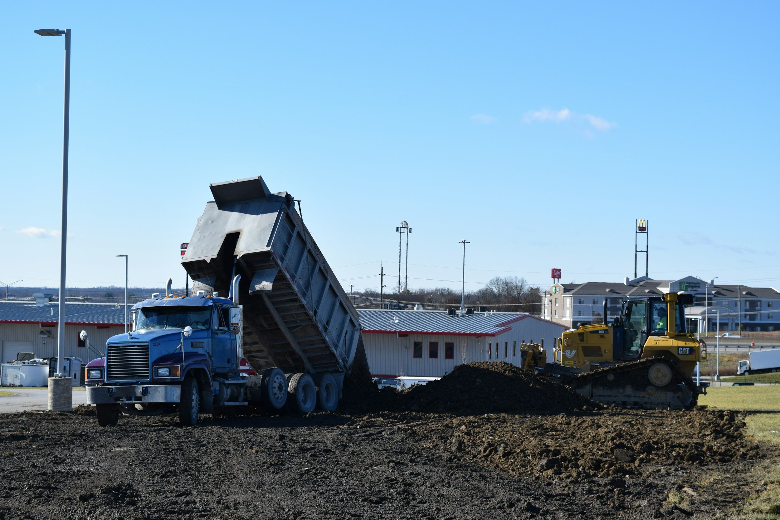 Construction site with a blue dump truck, a yellow bulldozer, and a building with a McDonald's sign in the background, under a clear blue sky.