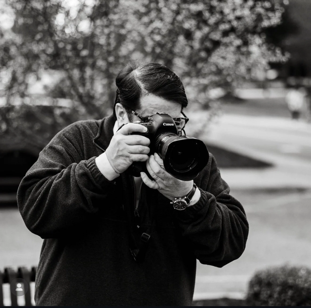 A man with dark hair taking a photograph outdoors with a camera, wearing glasses and a watch, in a park setting with trees in the background.