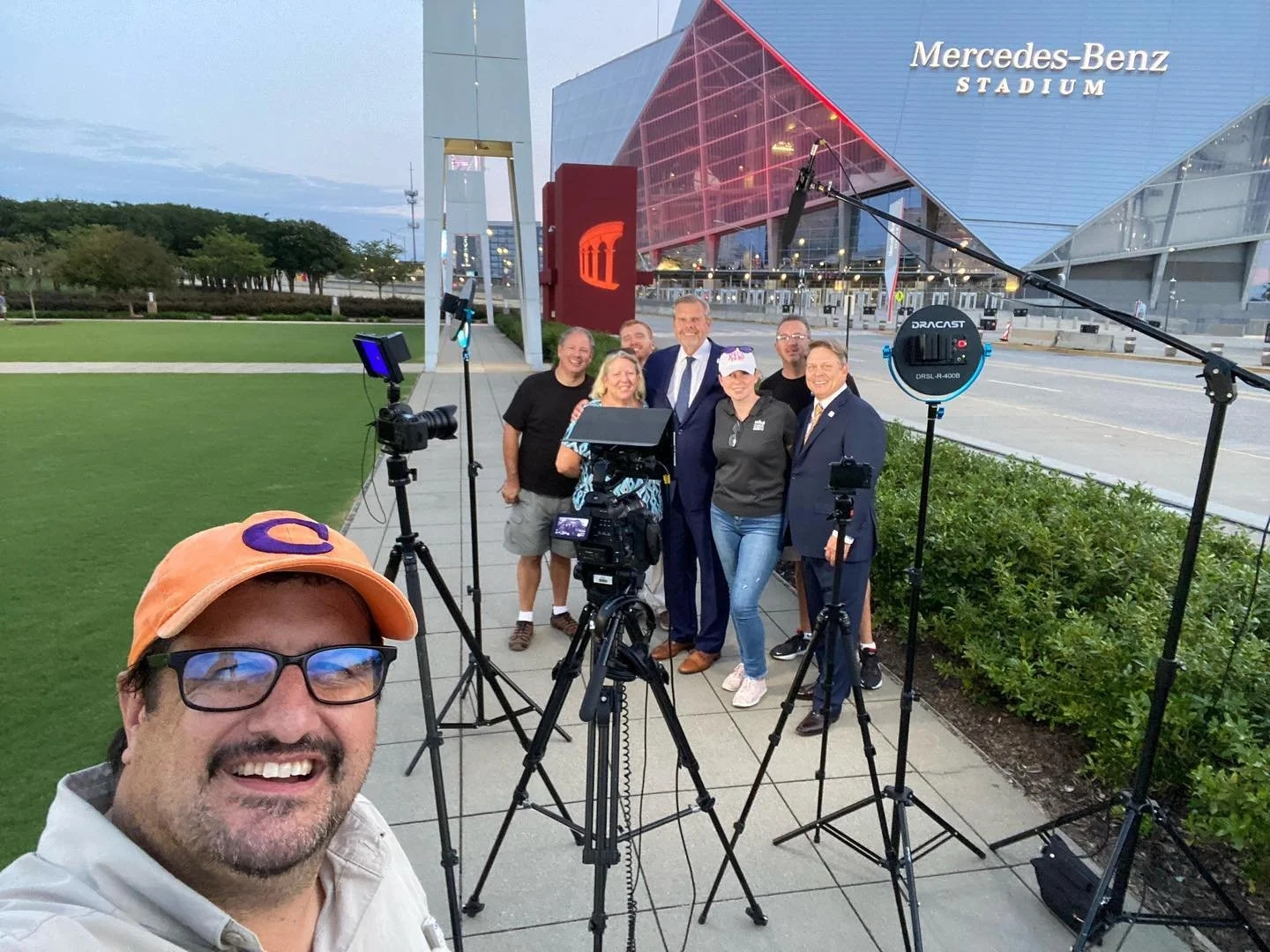 Group of people, including a man in a suit, standing outside Mercedes-Benz Stadium with cameras and filming equipment, taking a selfie with the stadium in the background during early evening.