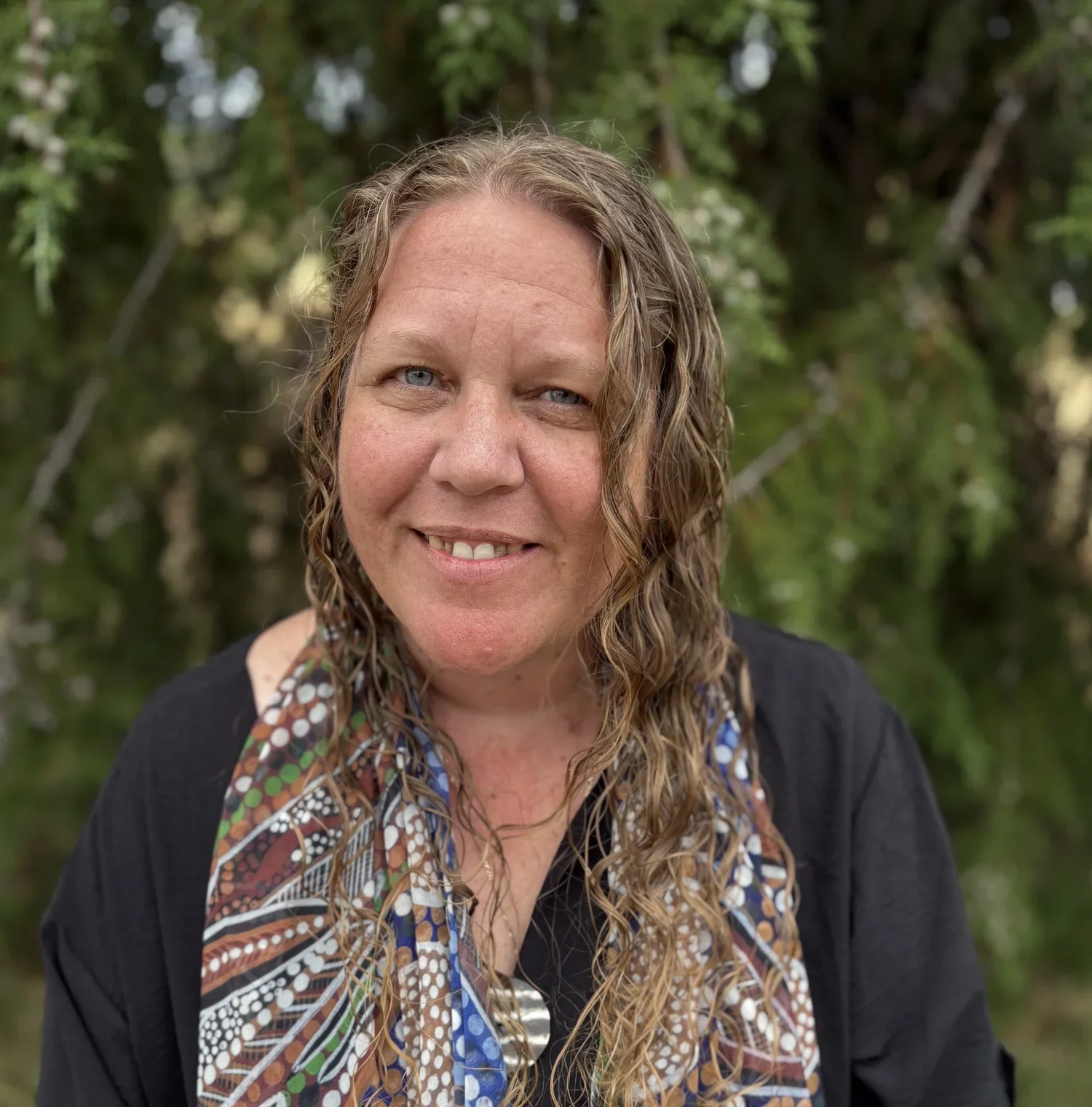 A woman with curly hair smiling outdoors with greenery in the background.