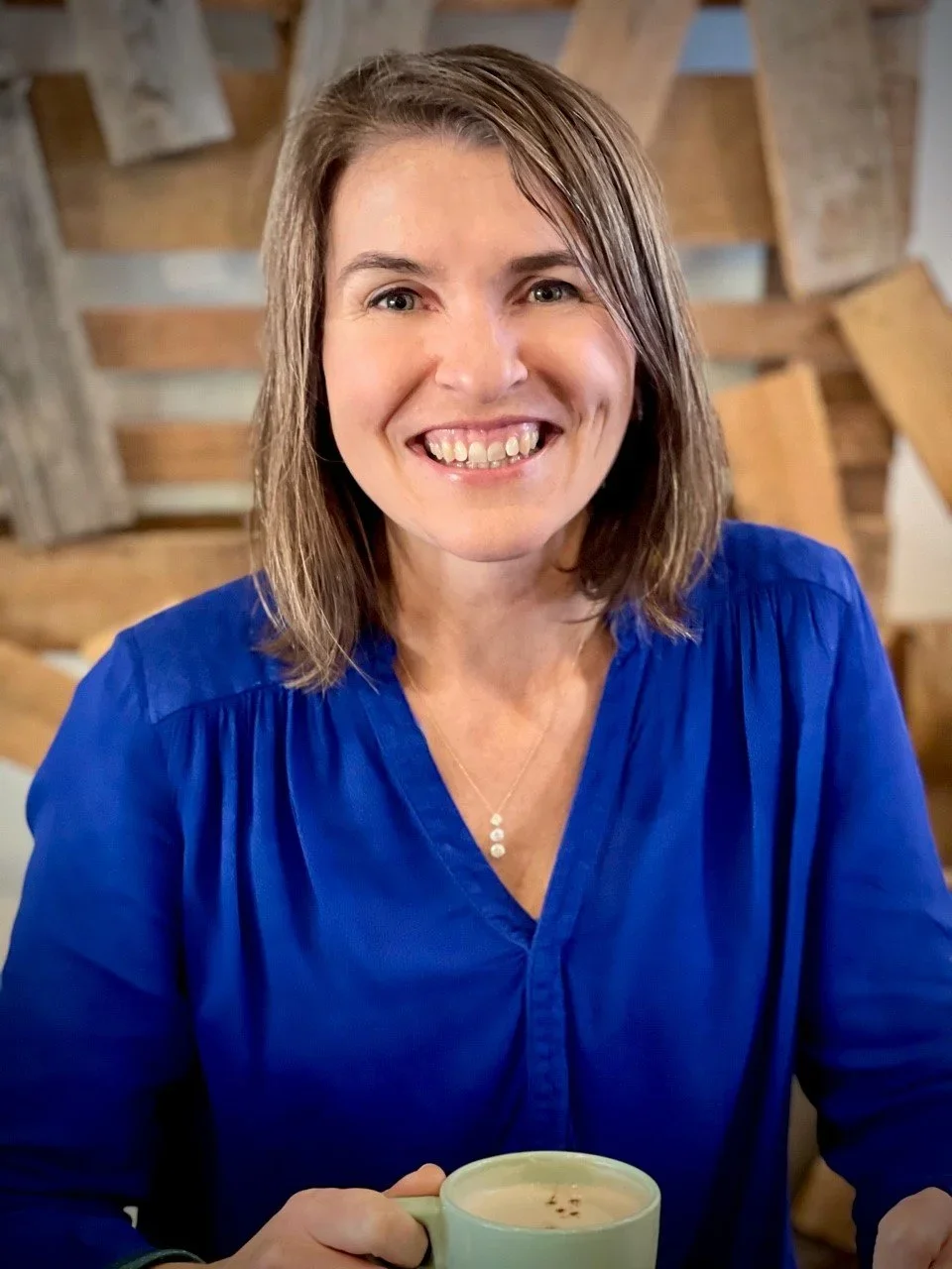 A woman with shoulder-length brown hair, smiling while holding a mug of coffee, wearing a blue blouse, sitting indoors with a wooden wall in the background.