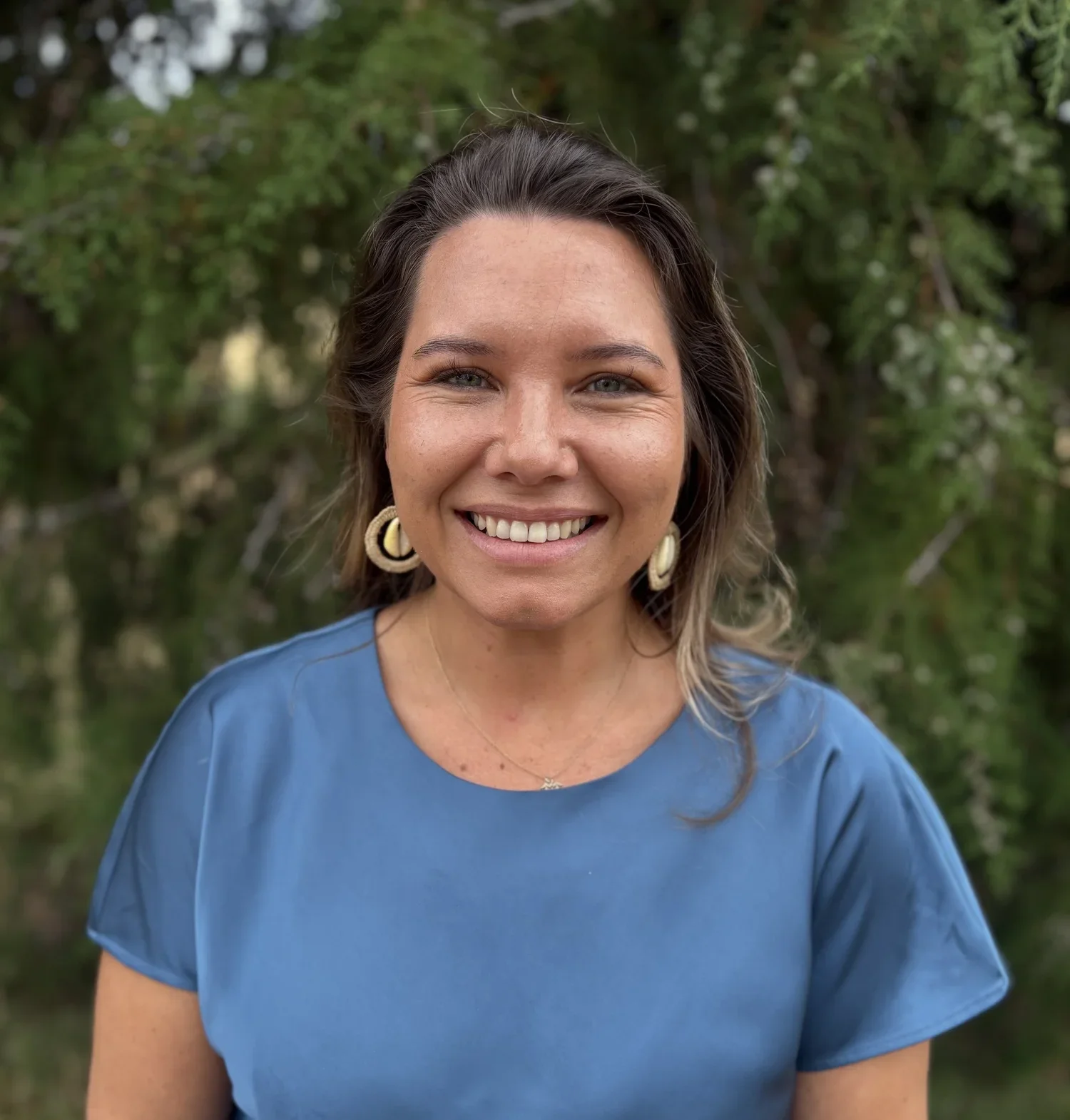 A smiling woman with wavy brown hair wearing a blue shirt and large earrings, standing outdoors with green trees in the background.