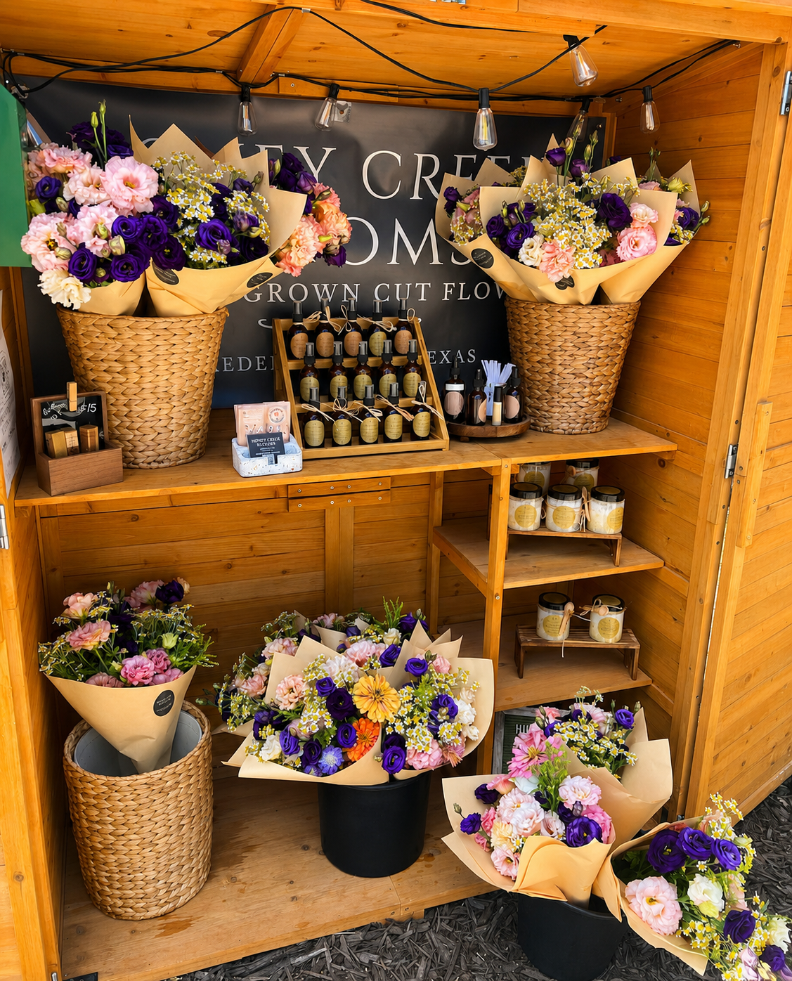 Market display of colorful flower bouquets in wicker baskets and black containers, with honey and candle products on wooden shelves, inside a wooden booth.