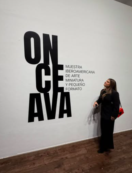 Gaby Rivera observando una pared con cartel que dice 'ONCE AVA' y texto adicional sobre la exhibición de arte miniatura y pequeño formato en Iberoamérica. donde Gaby participo y obtuvo reconocimiento.