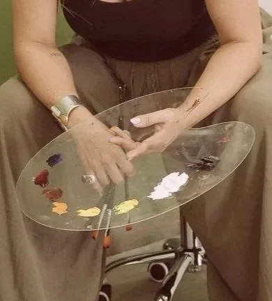 Persona sentada con pantalones beige, sosteniendo una paleta artística con manchas de pintura y una mano con manicura rosa, en un entorno de oficina o estudio.