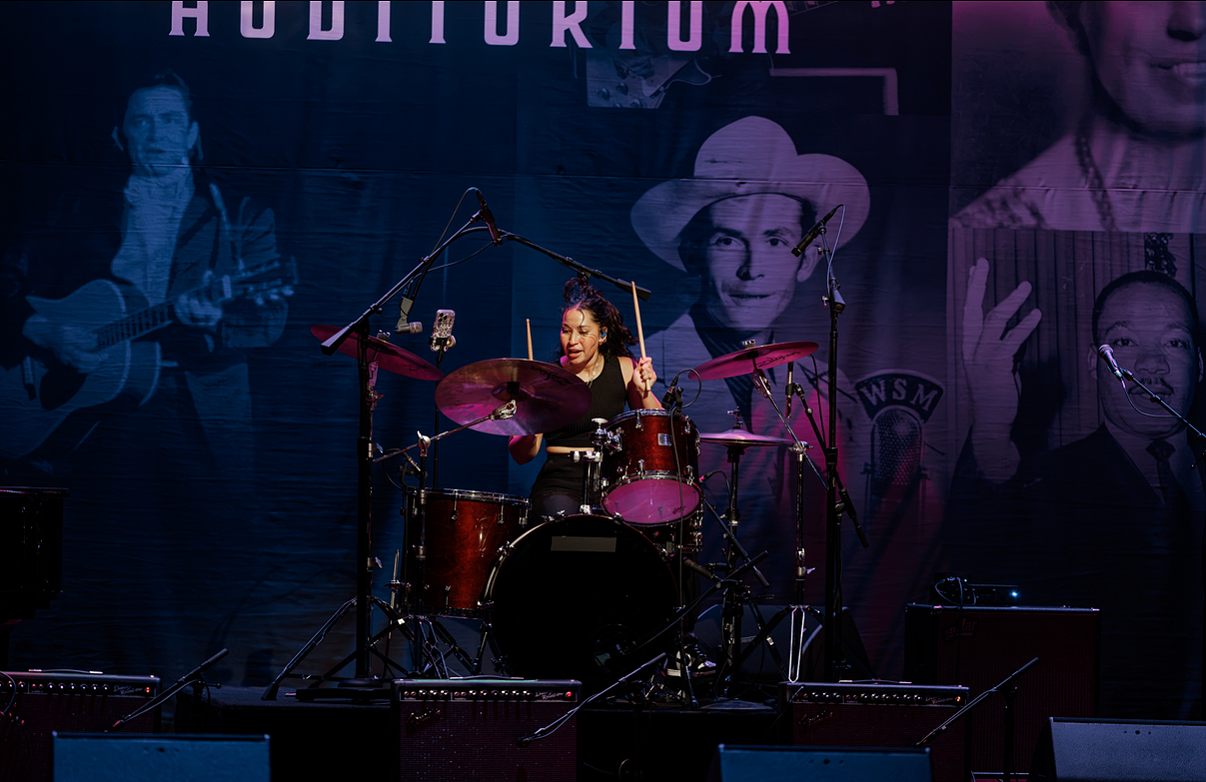 A woman playing drums on stage during a live music performance with large posters of musicians in the background.