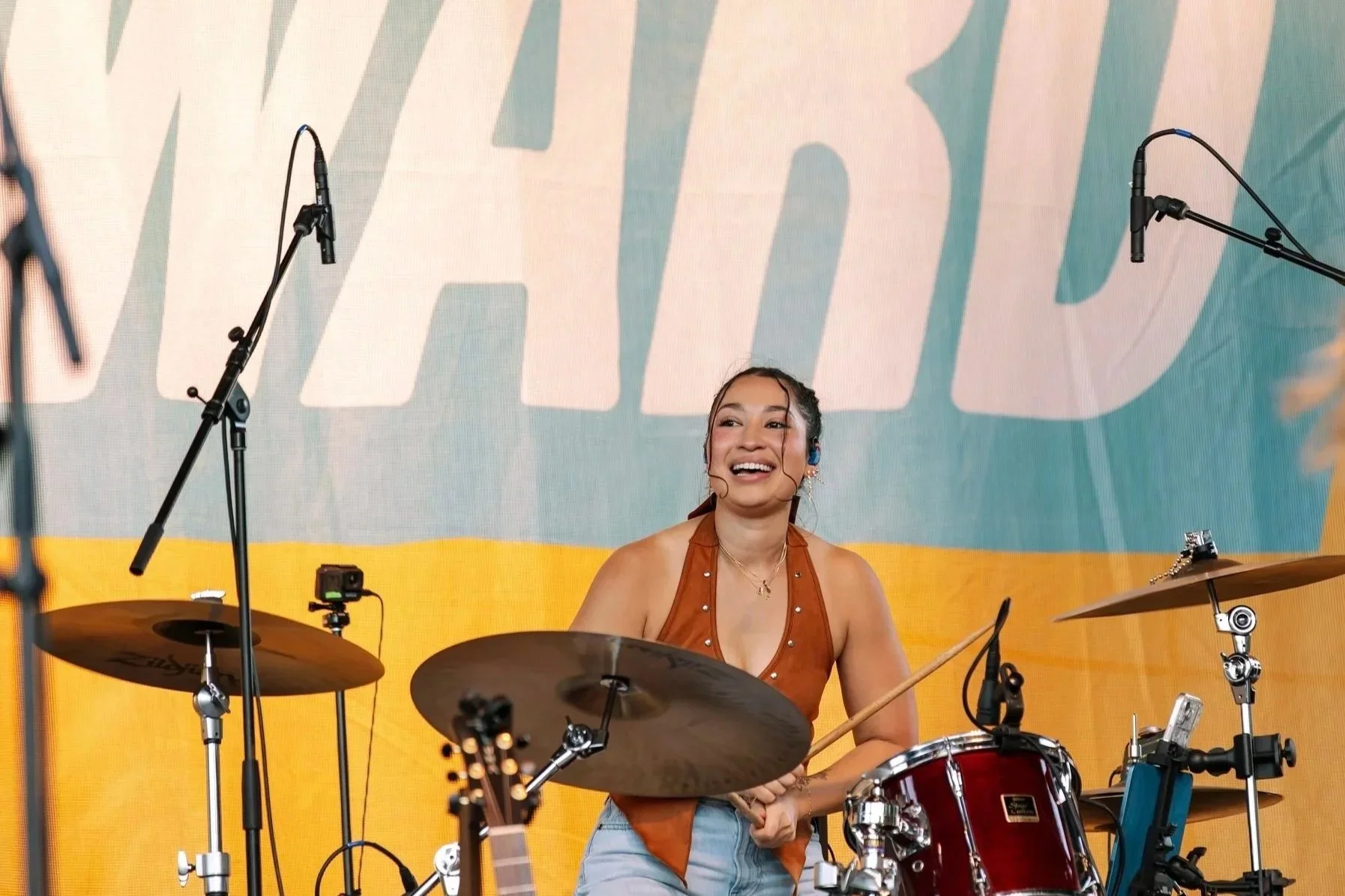A woman smiling and playing drums on stage with a colorful backdrop and microphones around her.