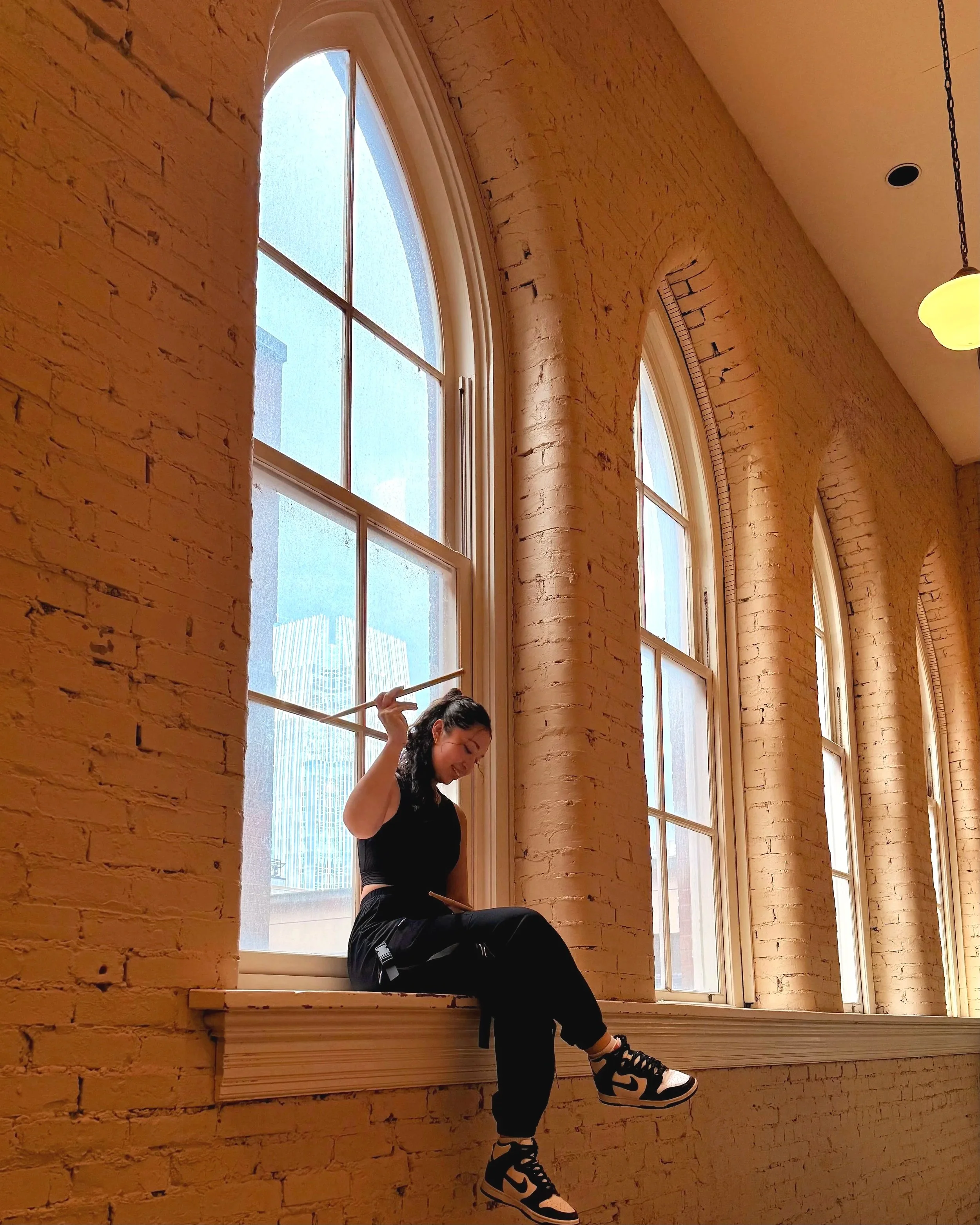 A young woman in black clothing and sneakers is sitting on a wooden window ledge, with her leg raised, in front of large arched windows in a room with brick walls and a warm glow from sunlight.