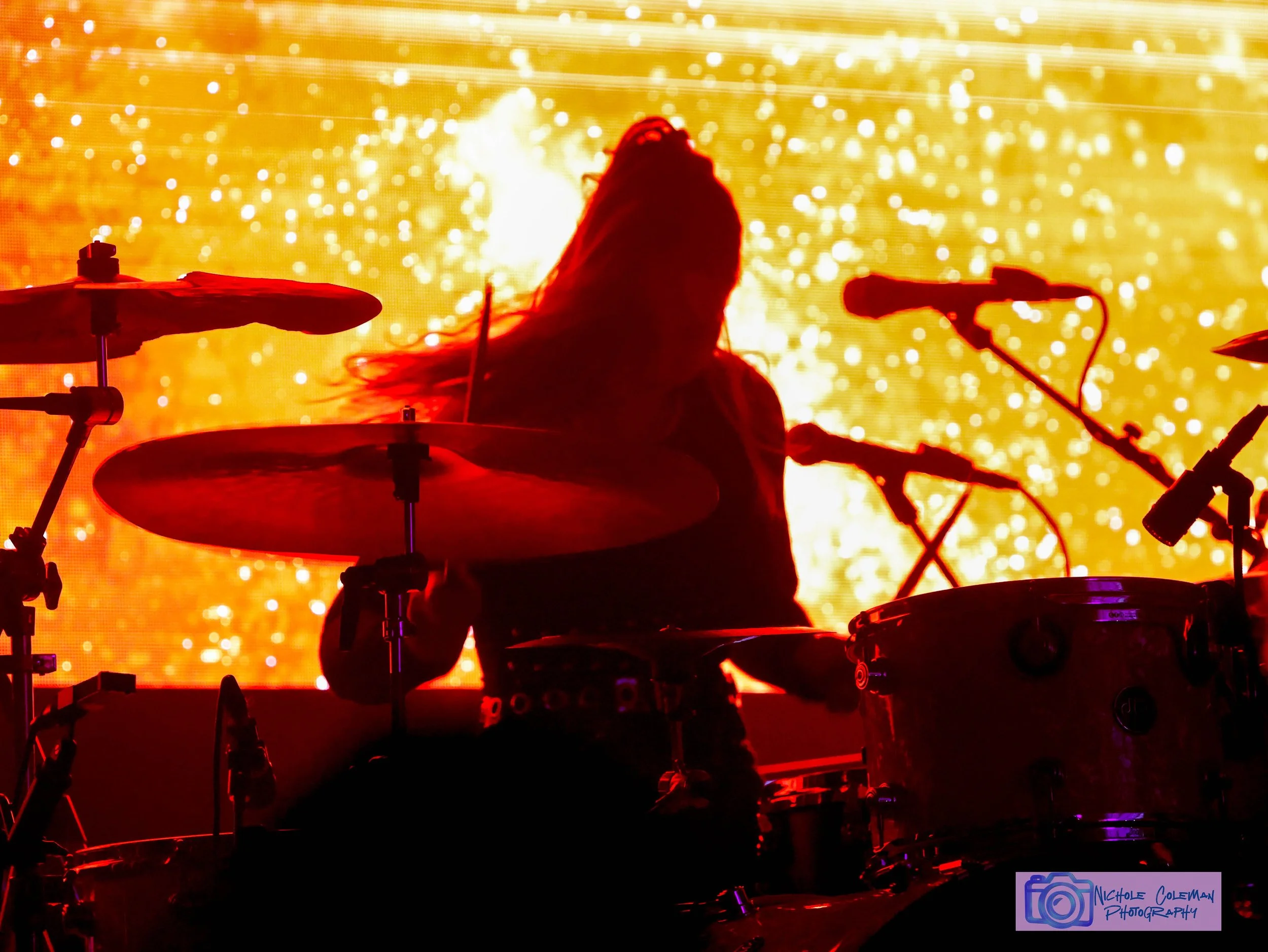 A drummer performing on stage with a vibrant yellow and orange background of bright lights and sparks, creating a dynamic concert scene.