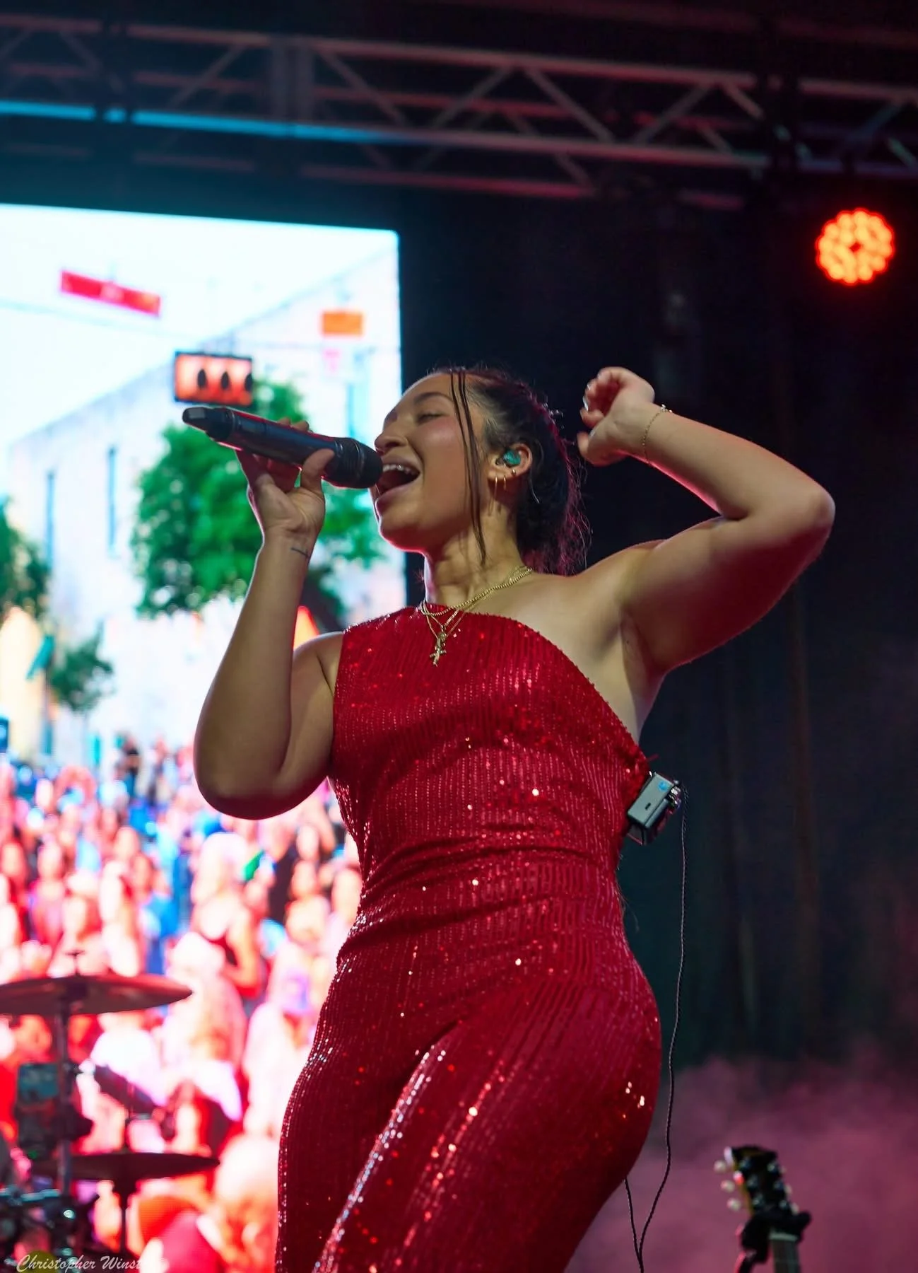 A woman singing into a microphone on stage, wearing a red, sequined dress with a necklace, with a live audience in the background.