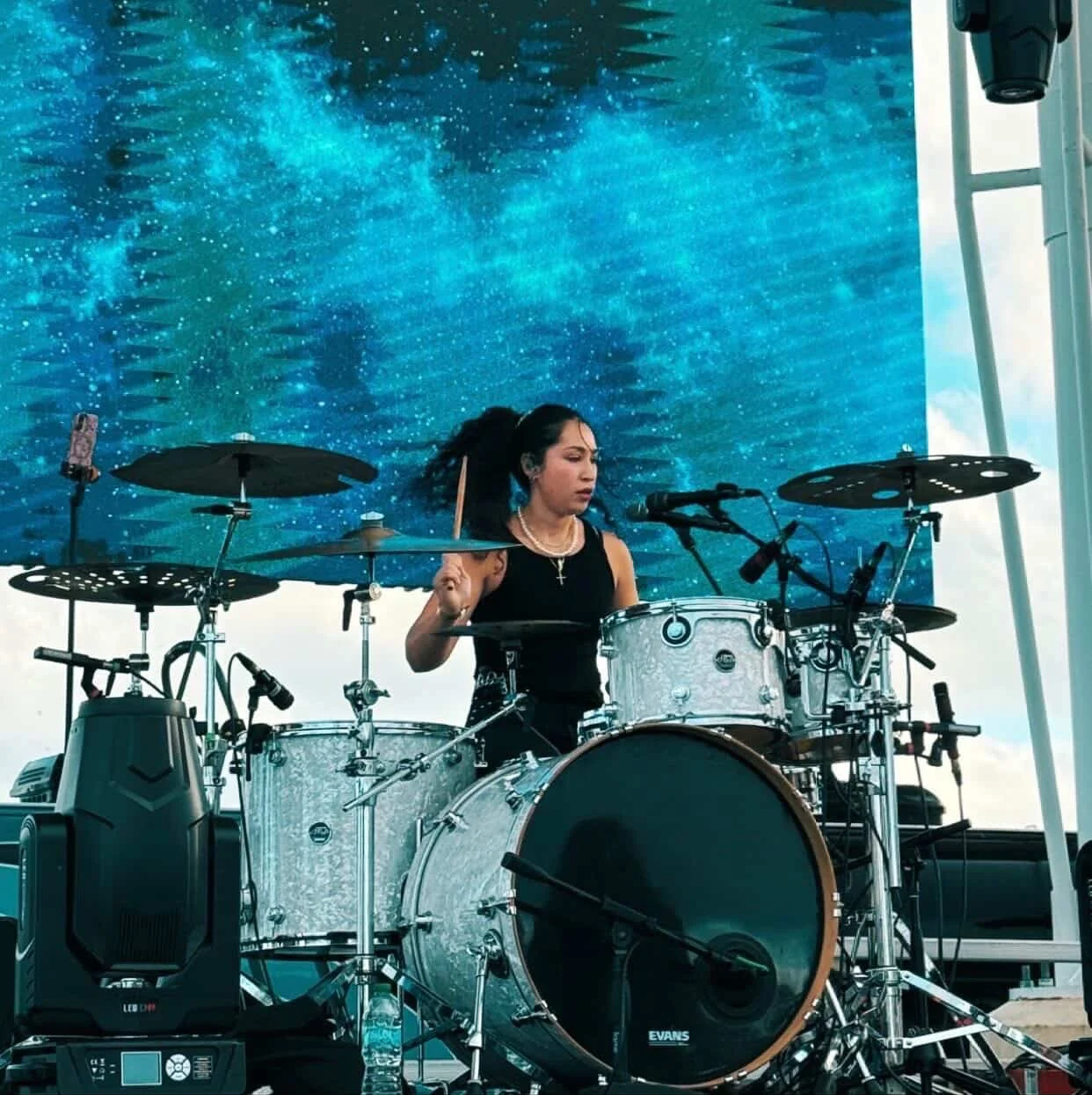 A woman playing a drum set on an outdoor stage with a blue sky background.