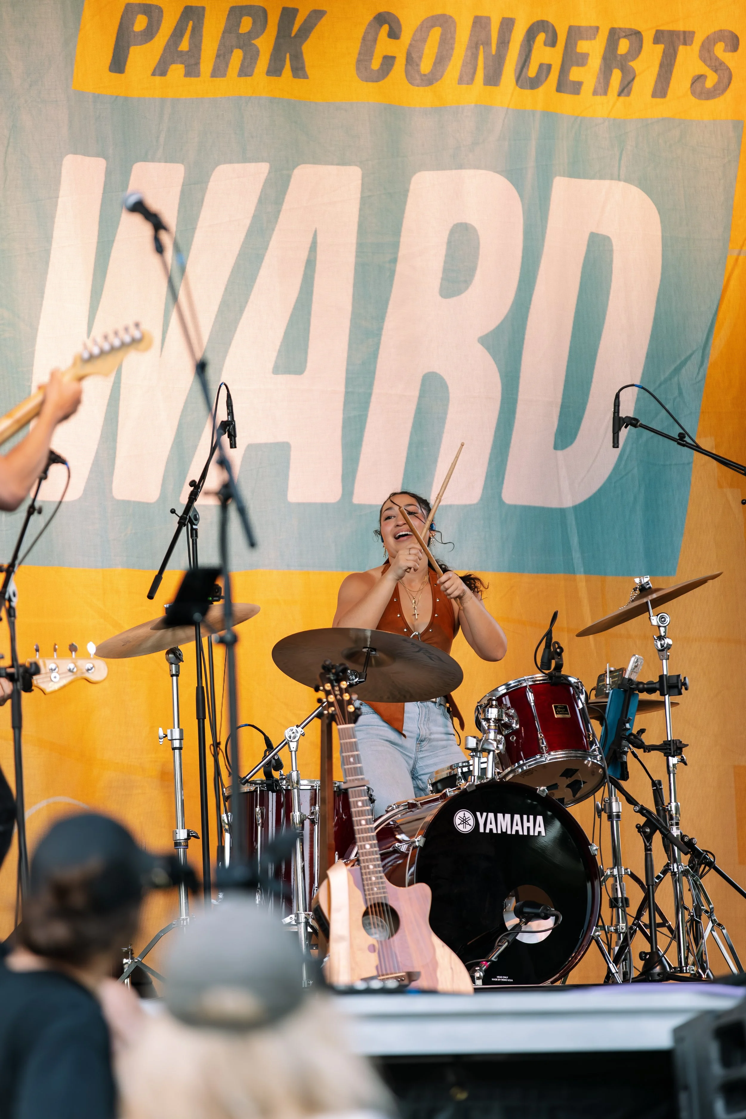 A woman is playing drums on a stage during a concert with a large yellow banner behind her that says 'PARK CONCERTS' and large, bold letters saying 'YARD'. Other musicians and equipment, including guitars and microphones, are also visible on the stage.
