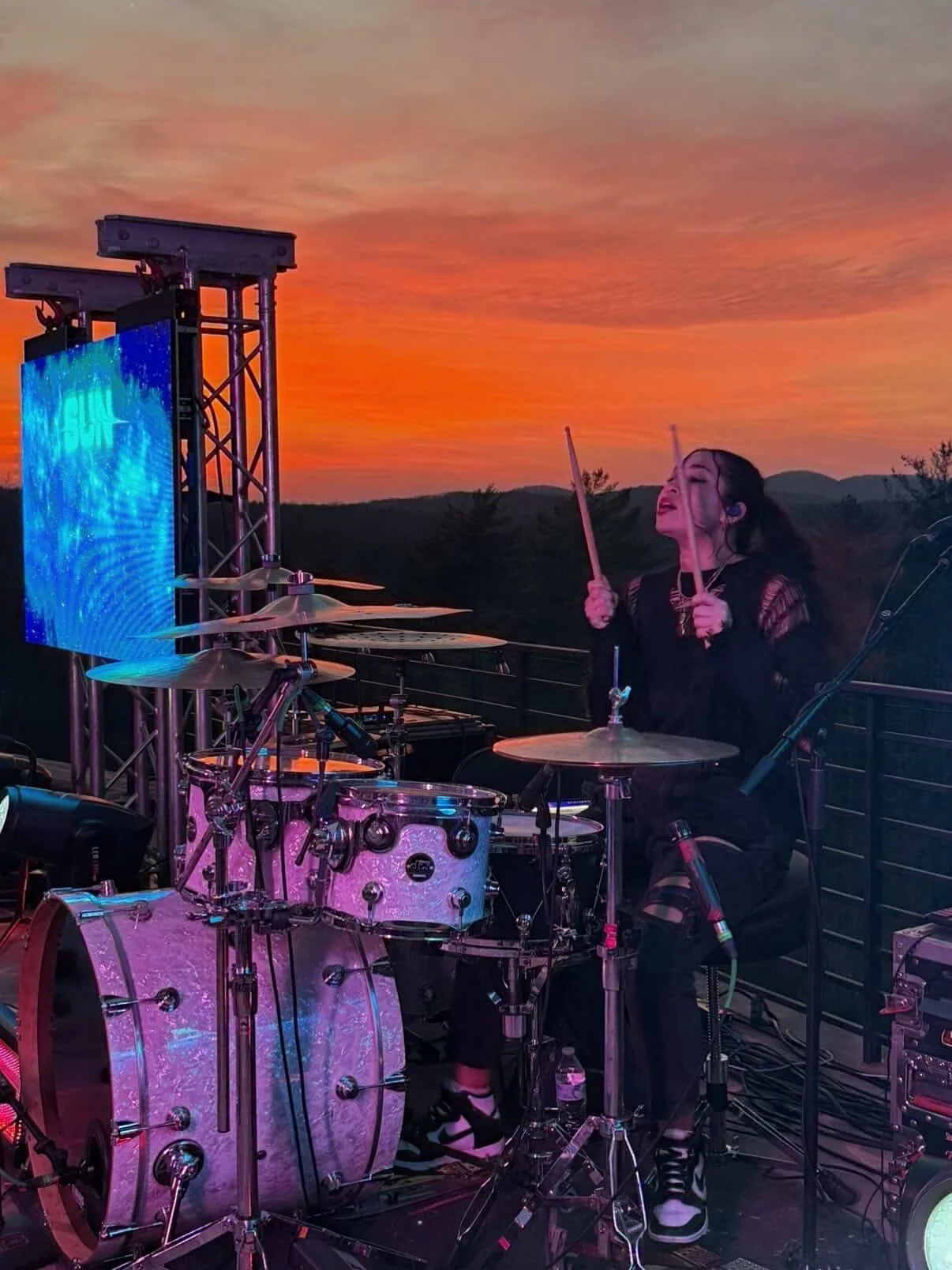 A female drummer playing drums on an outdoor stage during a sunset, with a large LED screen and sound equipment around her.