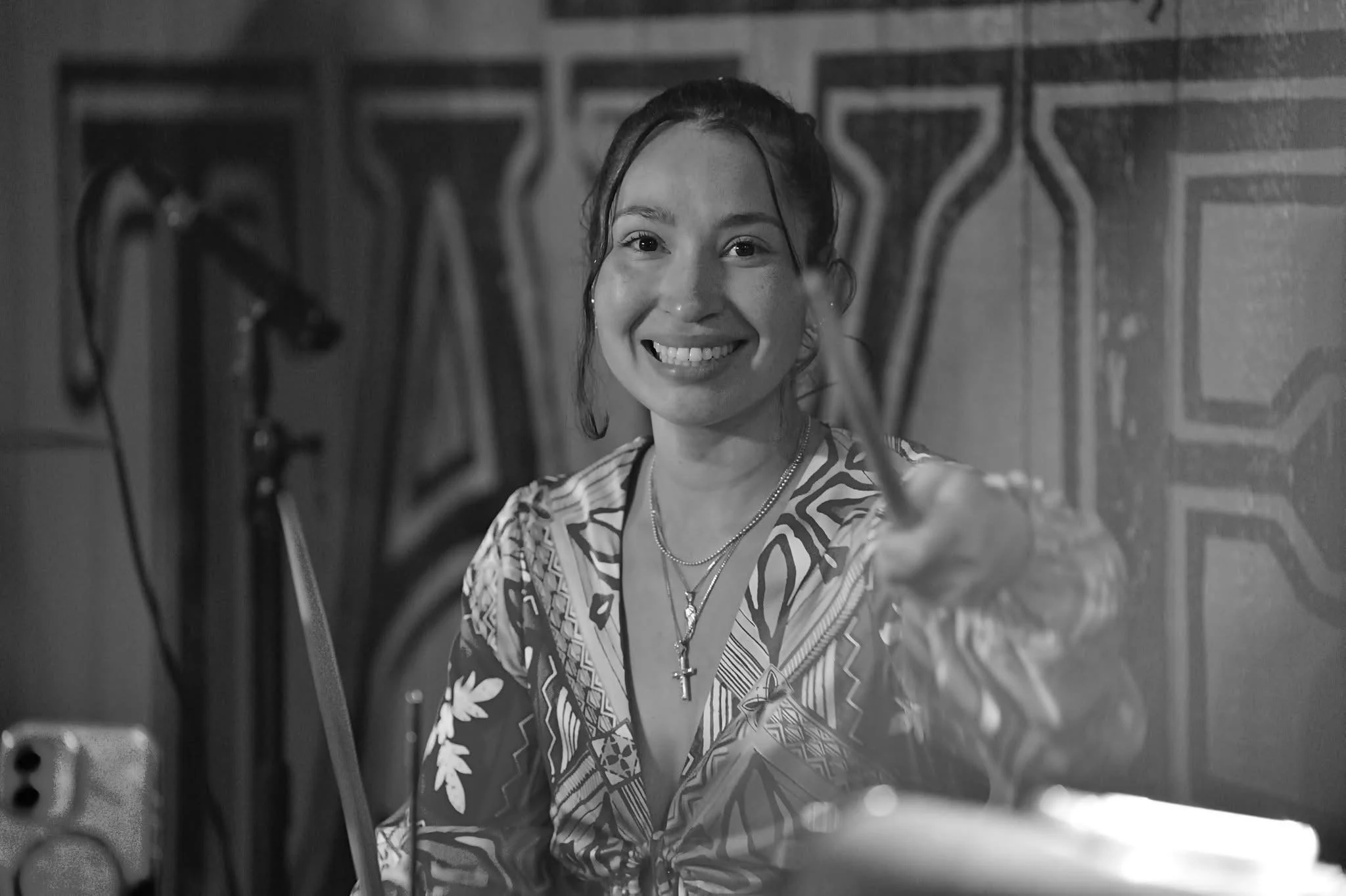 A smiling woman with dark hair, wearing a patterned shirt and layered necklaces, taking a selfie in front of a mirror, with grooming tools visible on a shelf in the background.