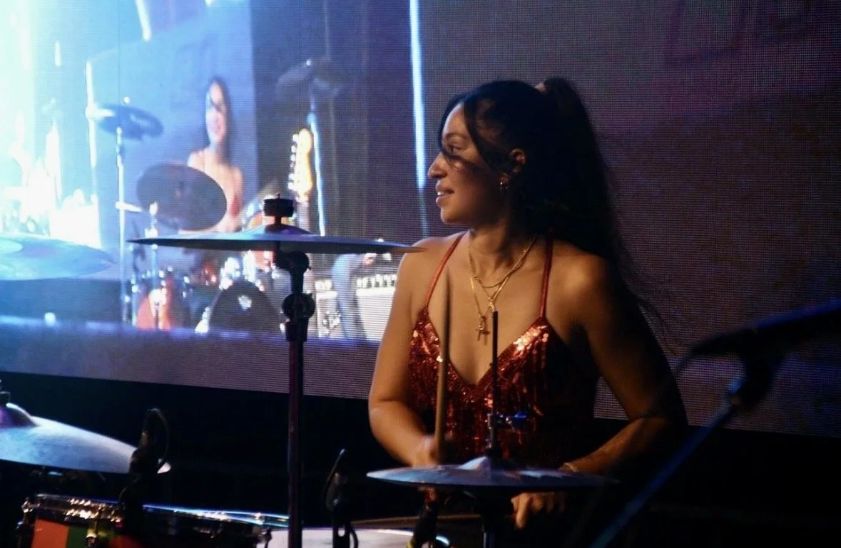 A woman playing drums at a music performance, wearing a red sequin top and gold jewelry, with a large screen showing her image in the background.