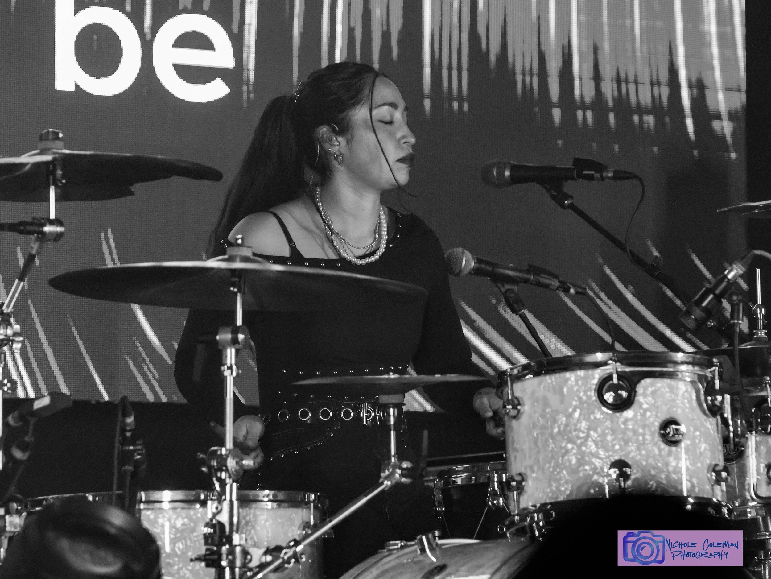 A woman with dark hair, multiple earrings, and layered necklaces is playing drums on stage with closed eyes, seen from the side amidst a set of drums and cymbals. In the background, there is a screen displaying partially visible text and graphics.