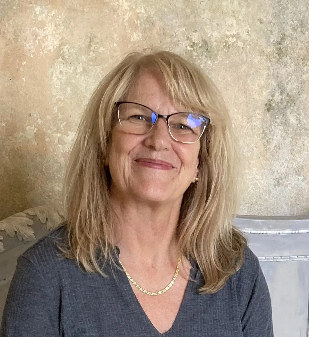 A middle-aged woman with blonde, shoulder-length hair and glasses, smiling at the camera, wearing a dark gray top and a gold necklace, standing in front of a textured beige wall.