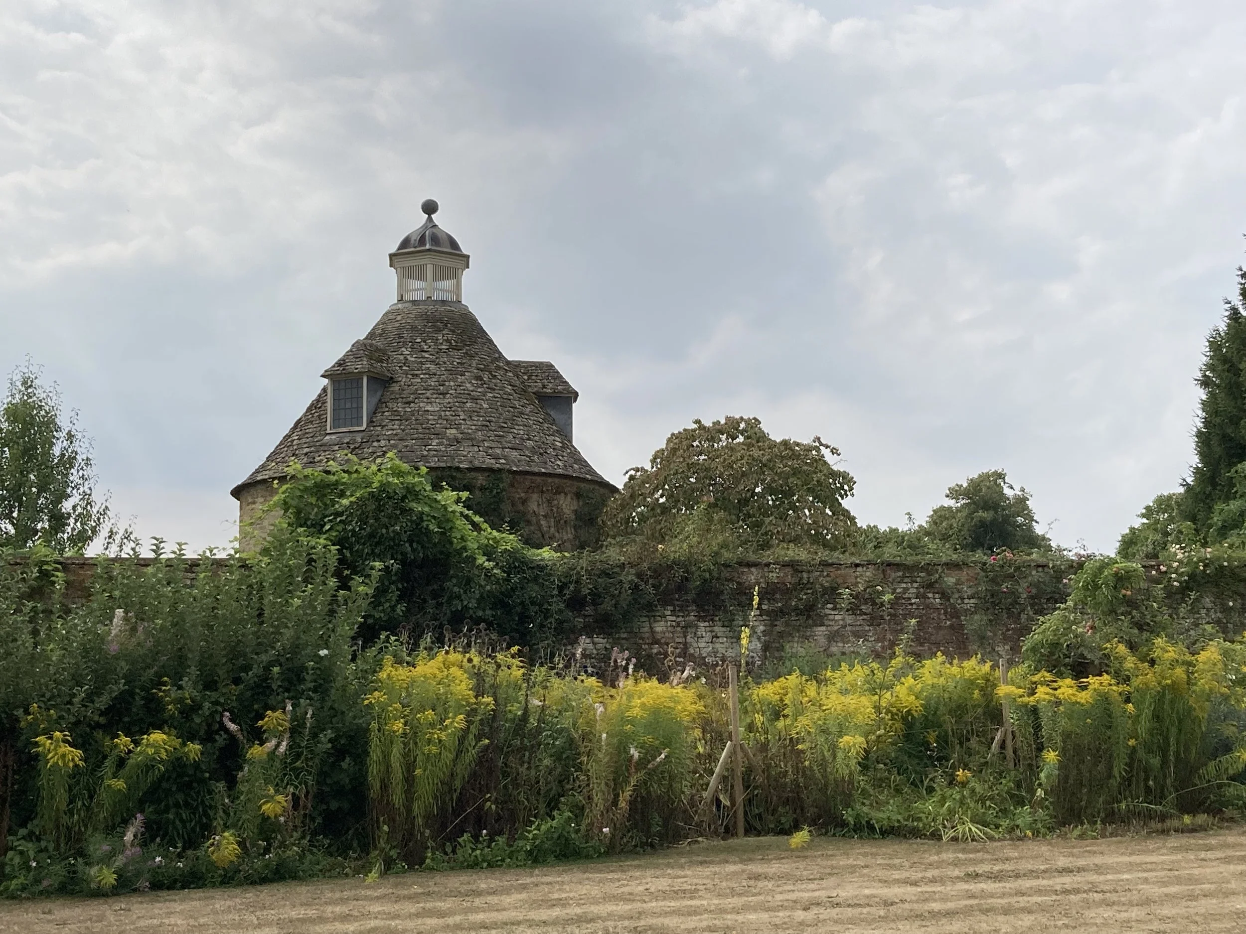A quaint building with a stone shingle roof and a small cupola on top, surrounded by lush green trees and colorful flowers, under a partly cloudy sky. Rousham Gardens Cotswolds England. British countryside
