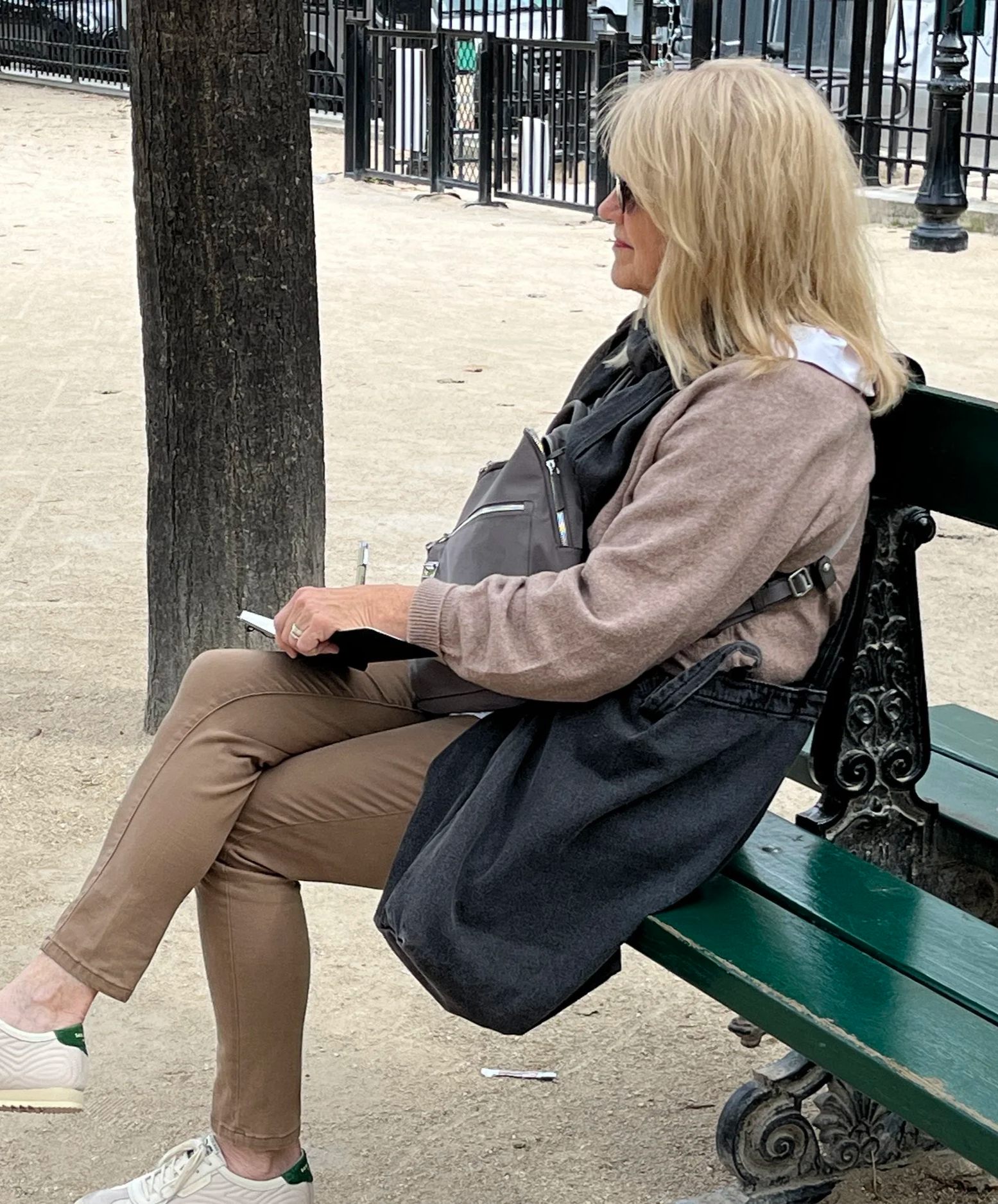 A woman with blonde hair wearing glasses, a beige sweater, and tan pants, sitting on a green park bench, holding a sketchbook in her right hand, drawing with a tree and a black wrought iron fence behind her.