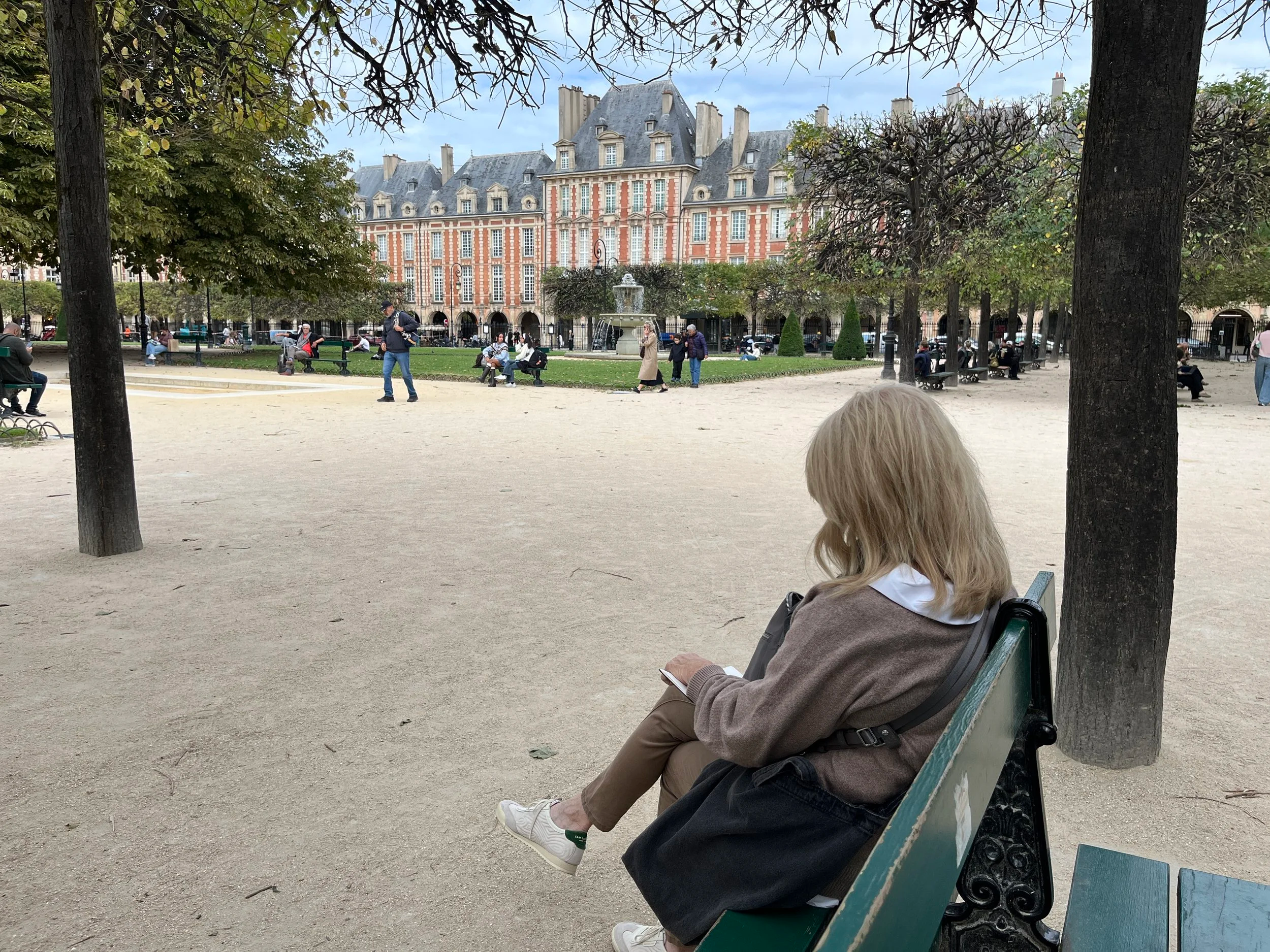 Woman with blonde hair sitting on a park bench, reading a book, with trees and a historic building in the background.