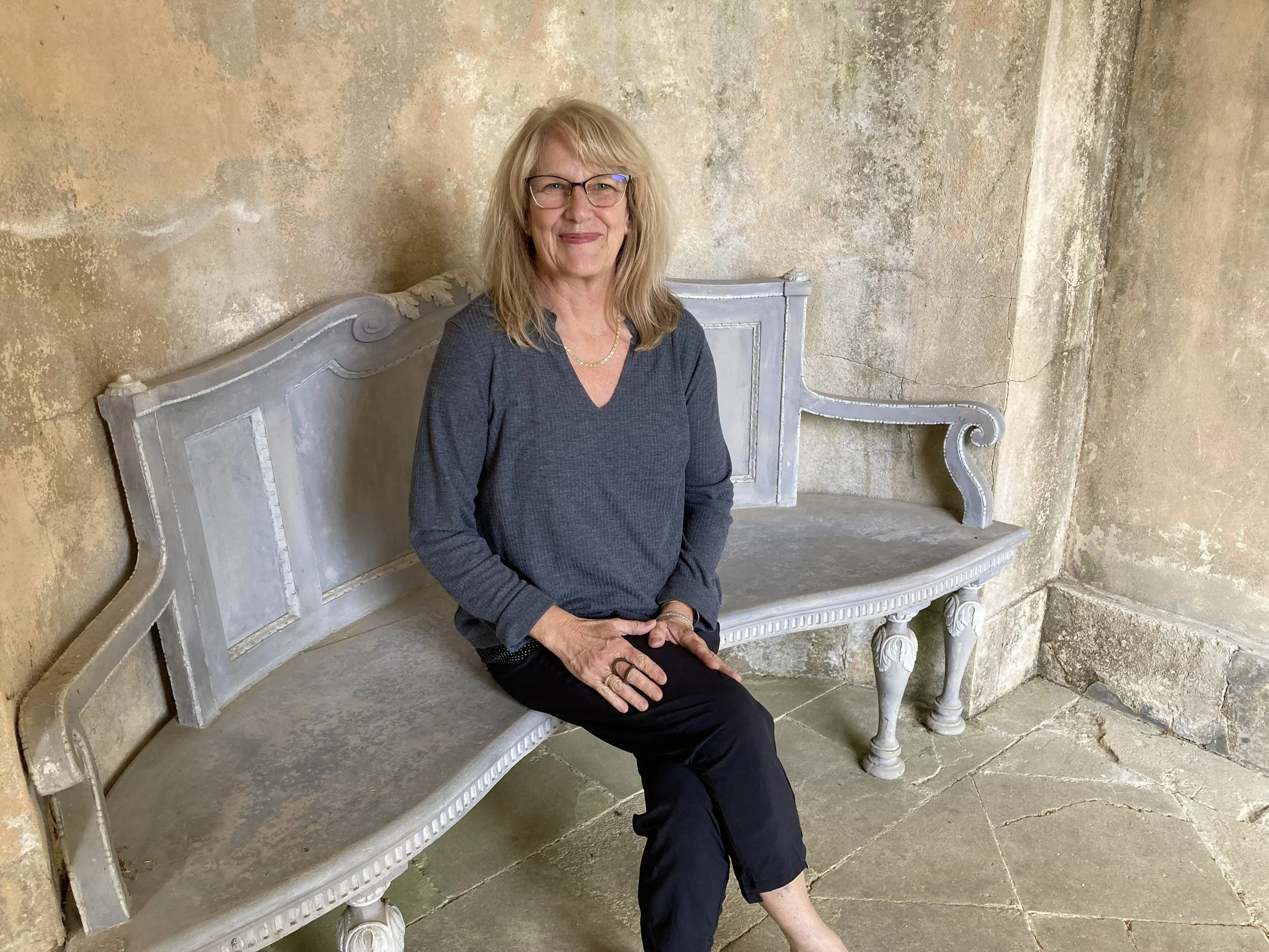 A smiling woman with blonde hair and glasses sits on a decorative shabby chic white bench against a weathered beige wall. Rousham Gardens, cotswold England  Brish countryside.