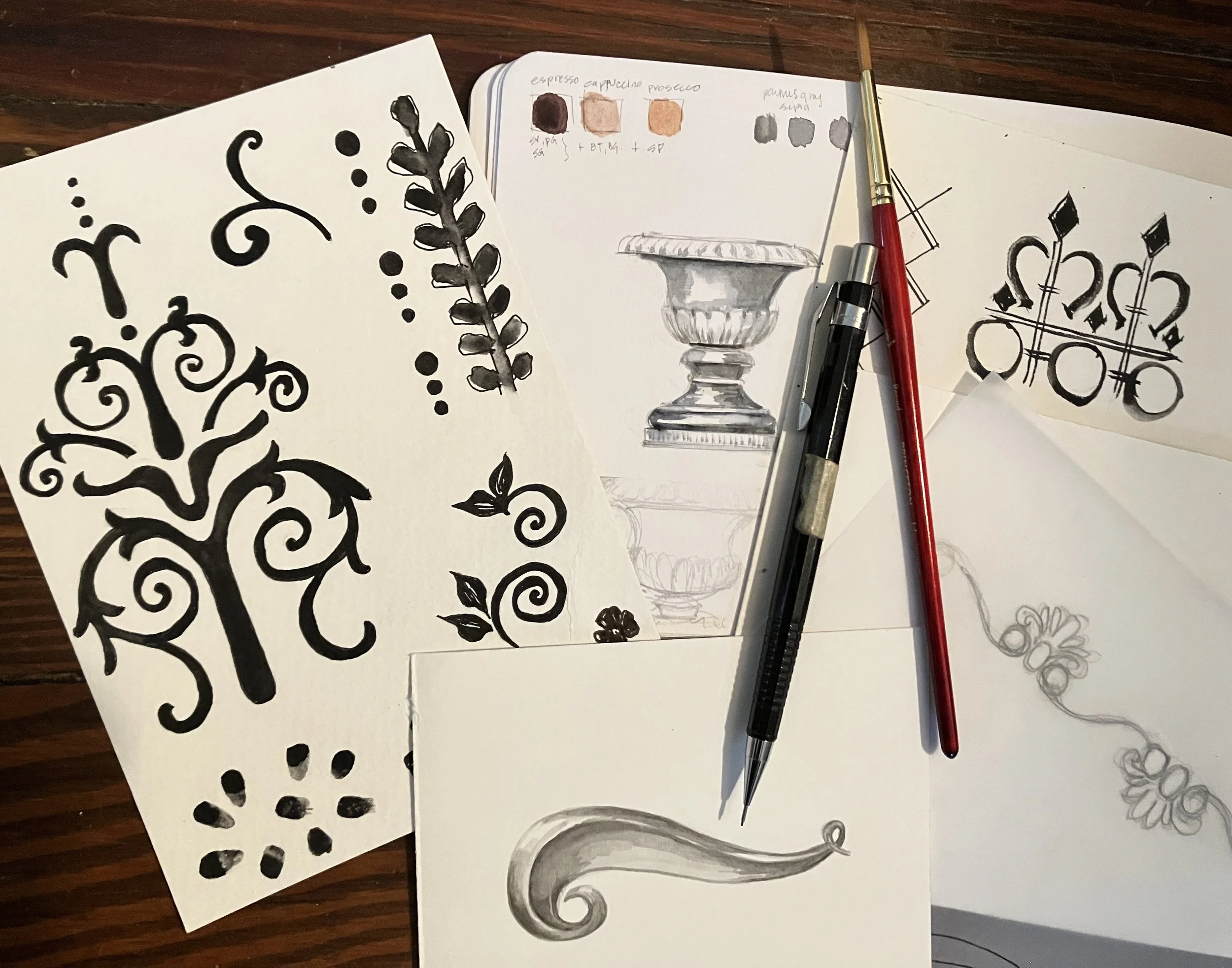 Several sheets of paper with black ink drawings, including decorative swirling designs and floral patterns. Two pens, one black and one red, rest on the papers. A watercolor swatch and a sketch of a pedestal vase are also visible.
