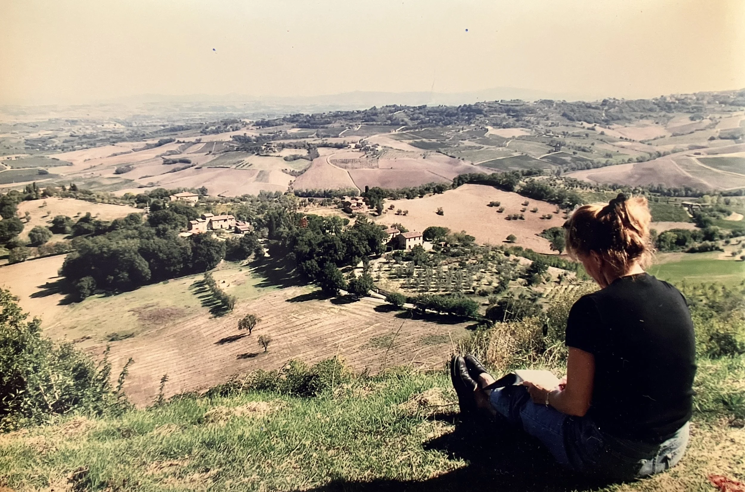 A person with long hair sitting on grass, looking at rolling hills and fields in the distance.  Montefollinico, Tuscany Italy