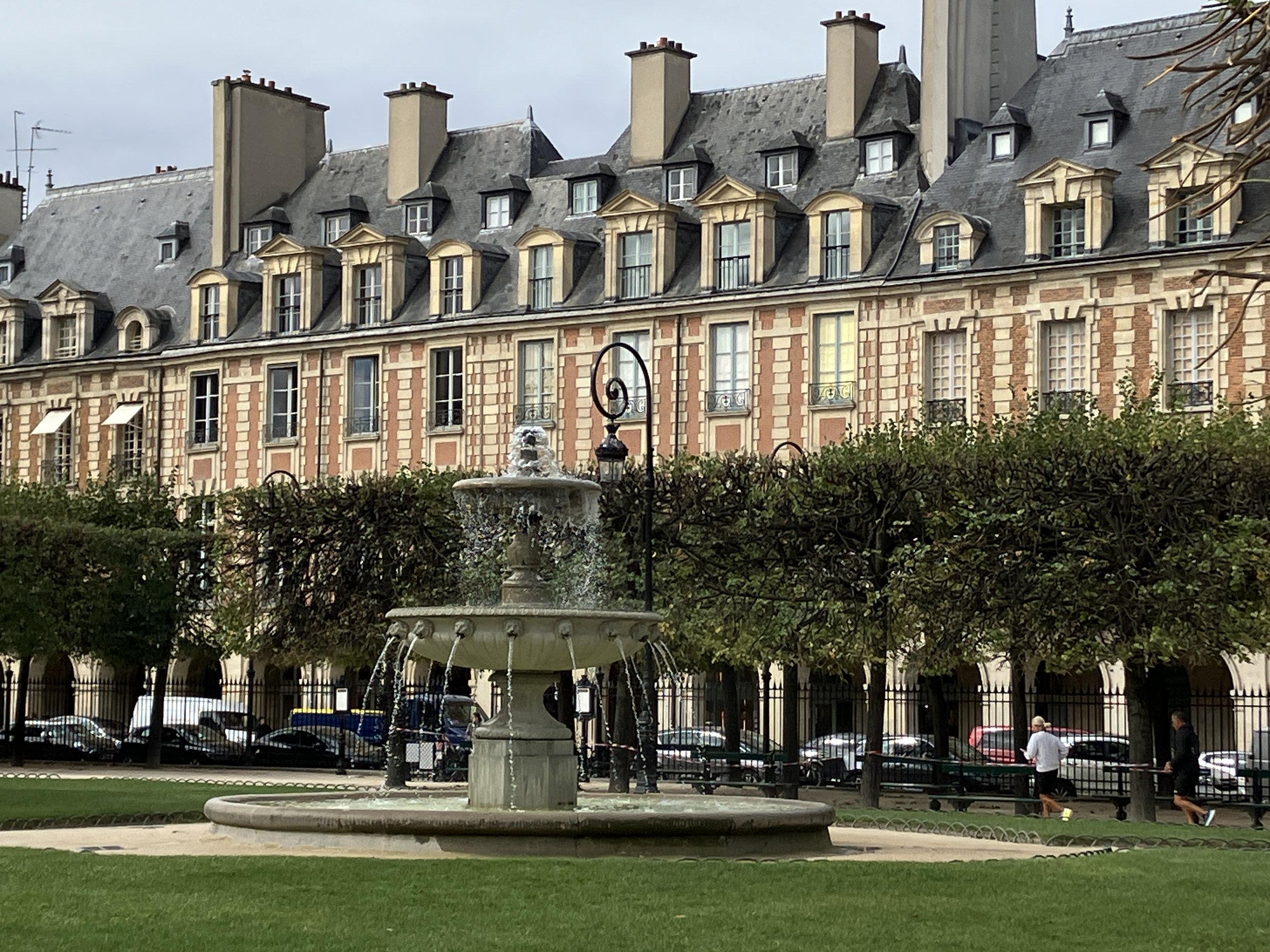 A decorative stone fountain with multiple water jets in a park surrounded by a low fence. In the background, there are ornate multi-story buildings with mansard roofs and dormer windows PLACE DES VOSGES Paris. People are walking/ sitting in the park.