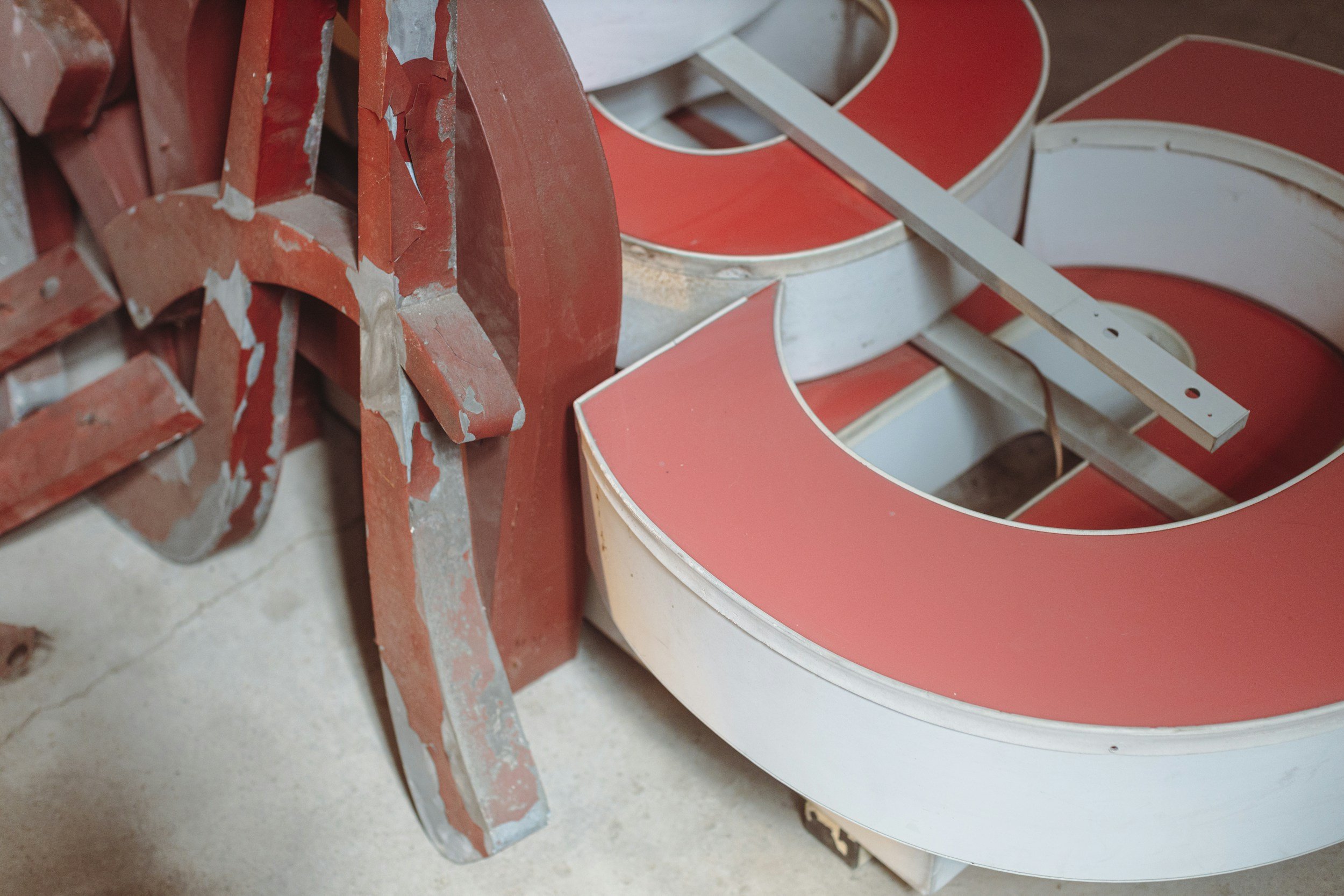 Close-up of a vintage, partially worn red and white neon sign with some peeling paint, resting on a concrete floor.