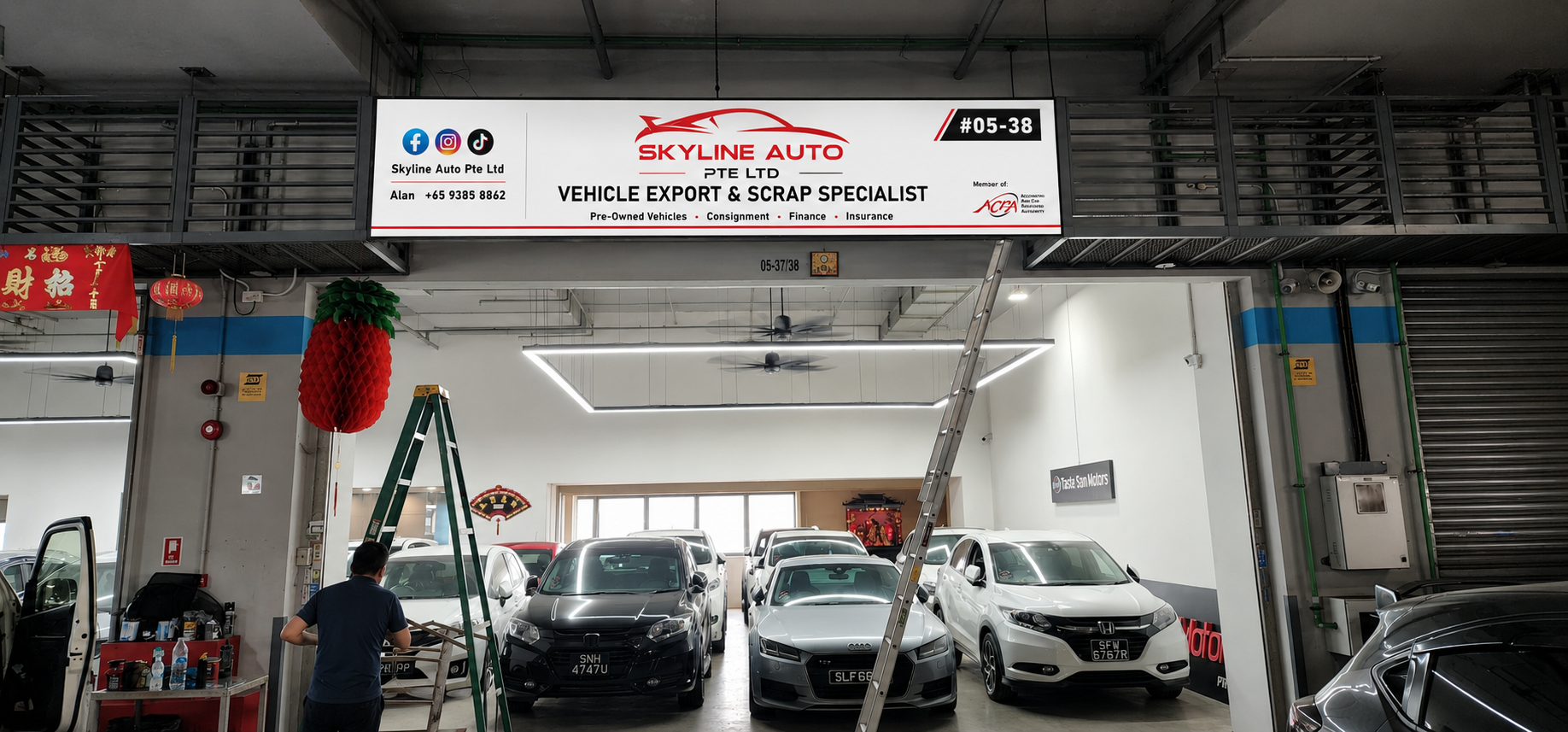 Indoor car dealership with multiple new cars parked inside. Sign overhead displaying information for Skyline Auto Pte Ltd, vehicle export and scrap specialist, with contact details and social media icons. Decorative red Chinese lanterns hanging inside.
