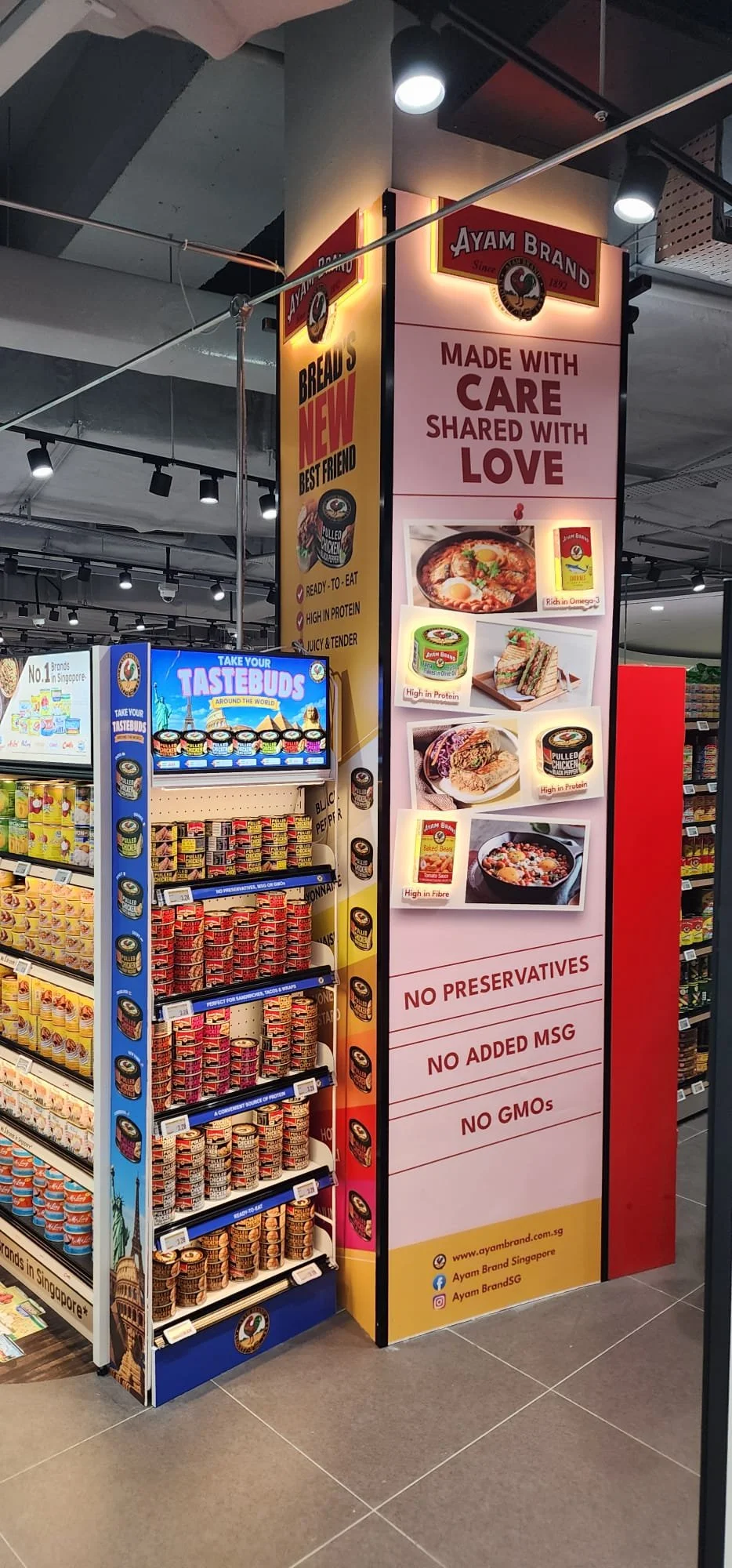 Promotional display for Ayam Brand with photographs of various food products, information about the brand, and claims about health benefits, along with text highlighting no preservatives, no added MSG, and no GMOs, in a retail store.
