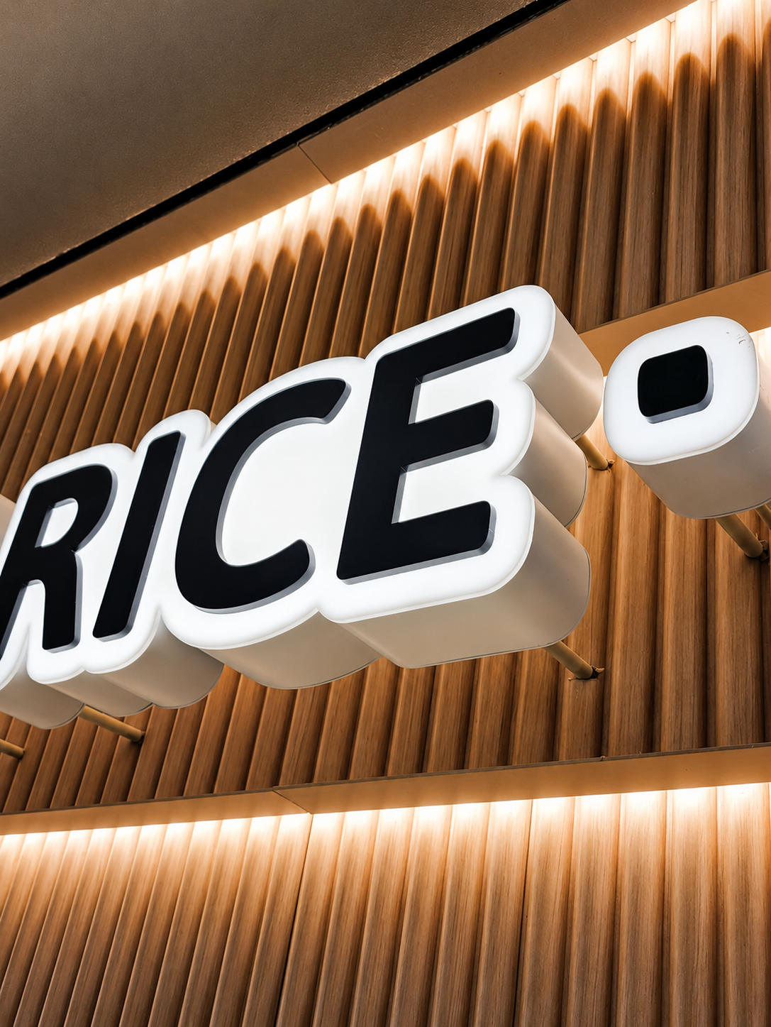Close-up of a illuminated sign with the partial word 'RICE' in black letters on a white background, mounted on a wooden slat wall.