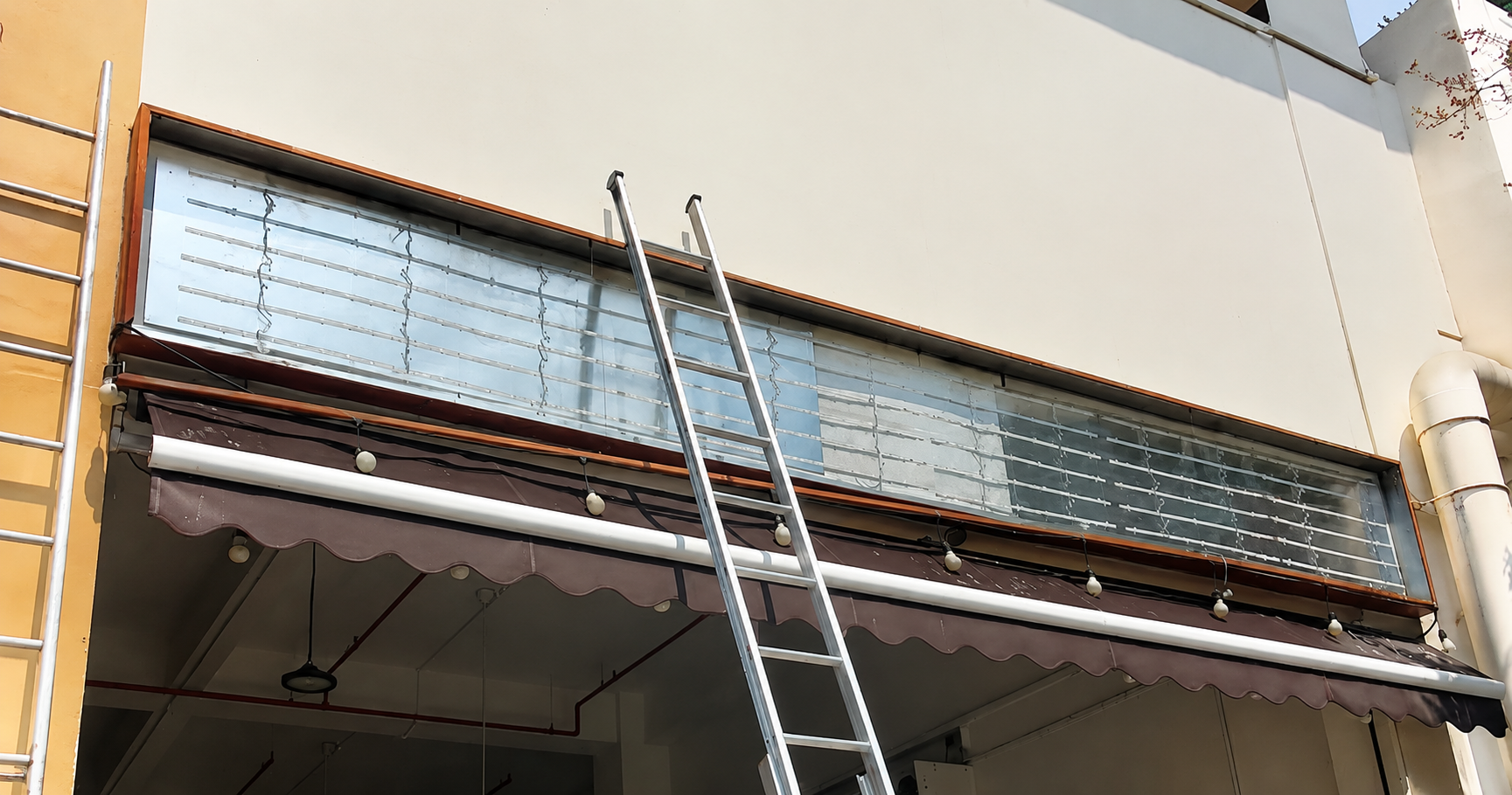 Building with a glass window, ladder leaning against it, and a brown canopy with string lights underneath.