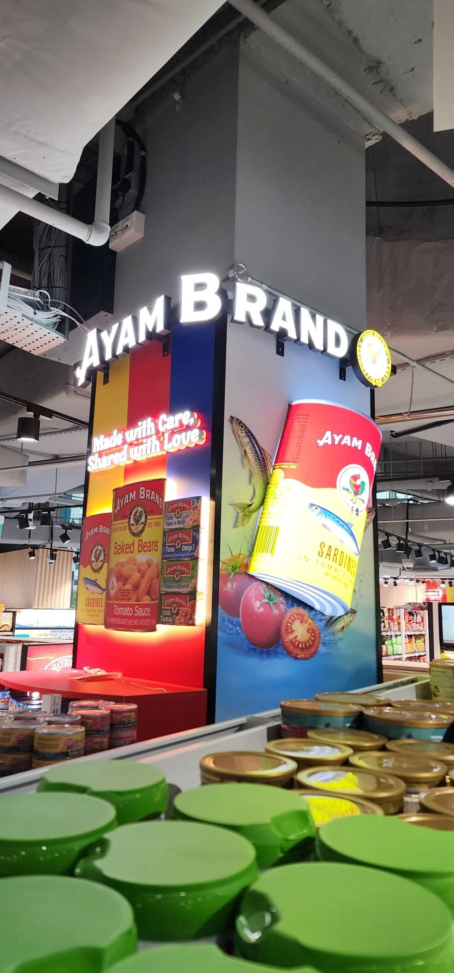 A display in a store featuring Ayam Brand canned foods, including baked beans and sardines, with signage that reads "Made with Care, Shared with Love" and colorful product packaging.