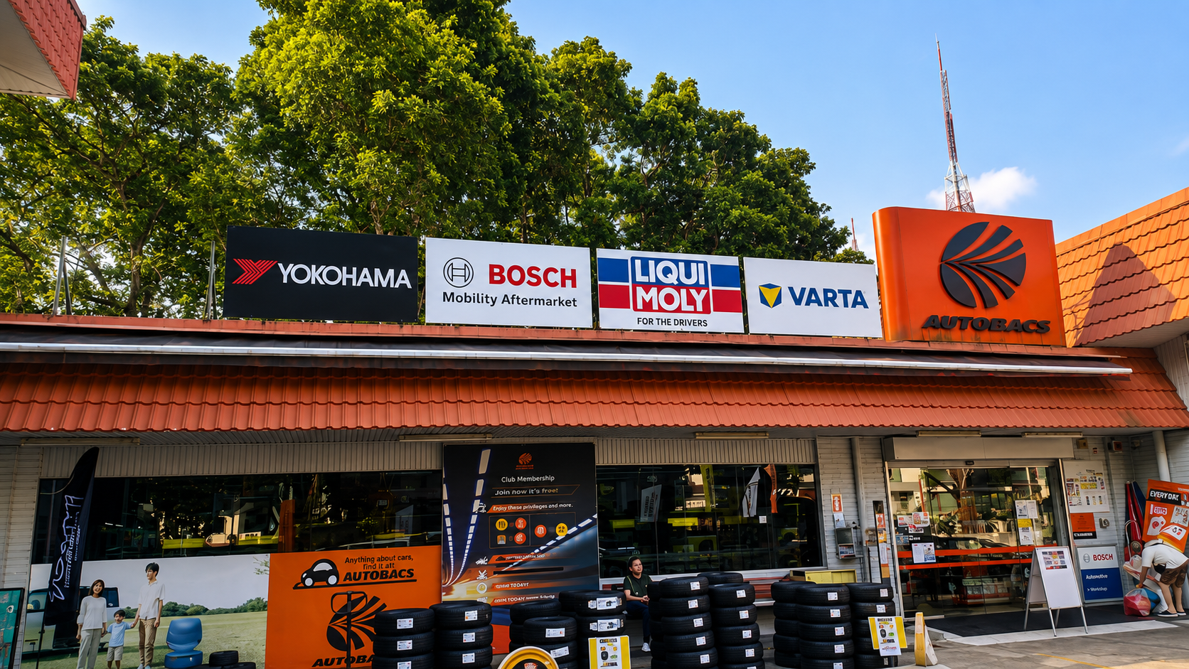 Exterior of an automotive store with signs from Yokohama, Bosch, Liqui Moly, Varta, and Autobacs. The store displays tires in front and has promotional banners and a few customers.