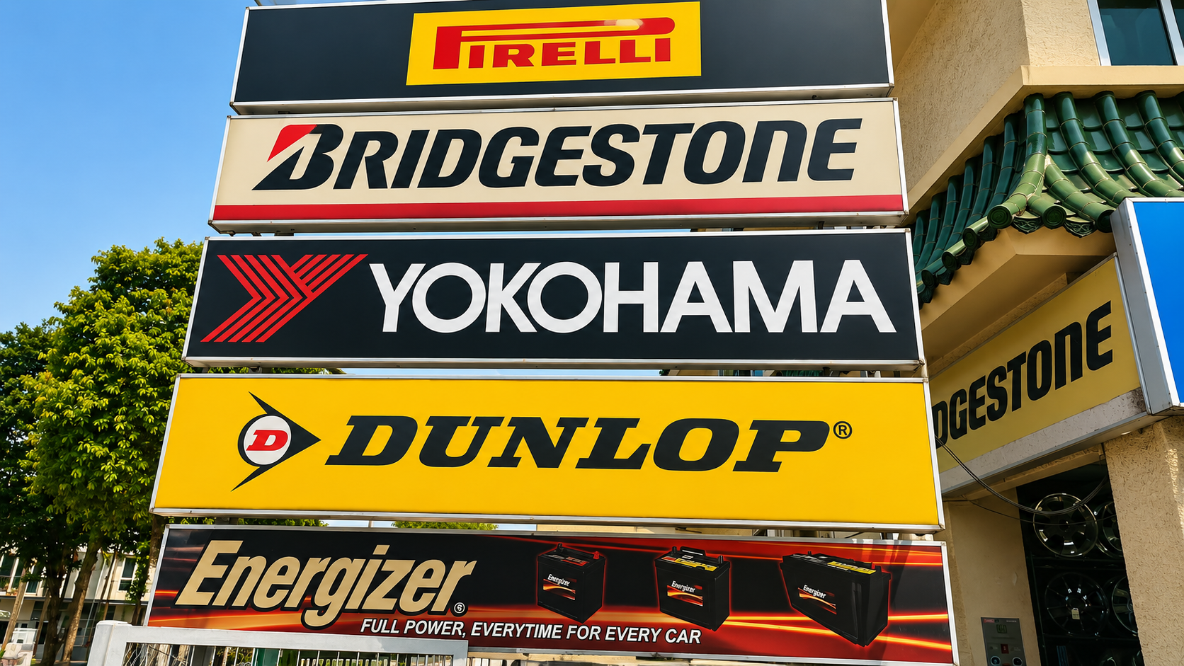 Signage for automotive brands Pirelli, Bridgestone, Yokohama, Dunlop, and Energizer in front of a building with green trees and blue sky.
