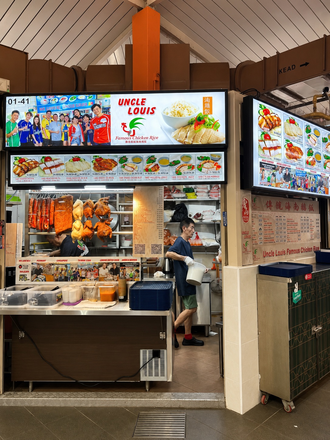 Food stall named Uncle Louis offering various dishes including roasted meats, chicken rice, and noodle dishes. Signs display images of menu items and a group of people at the top left. A man is working behind the counter, preparing food. Hanging roasted ducks and chickens are visible, along with containers of sauces on the counter.