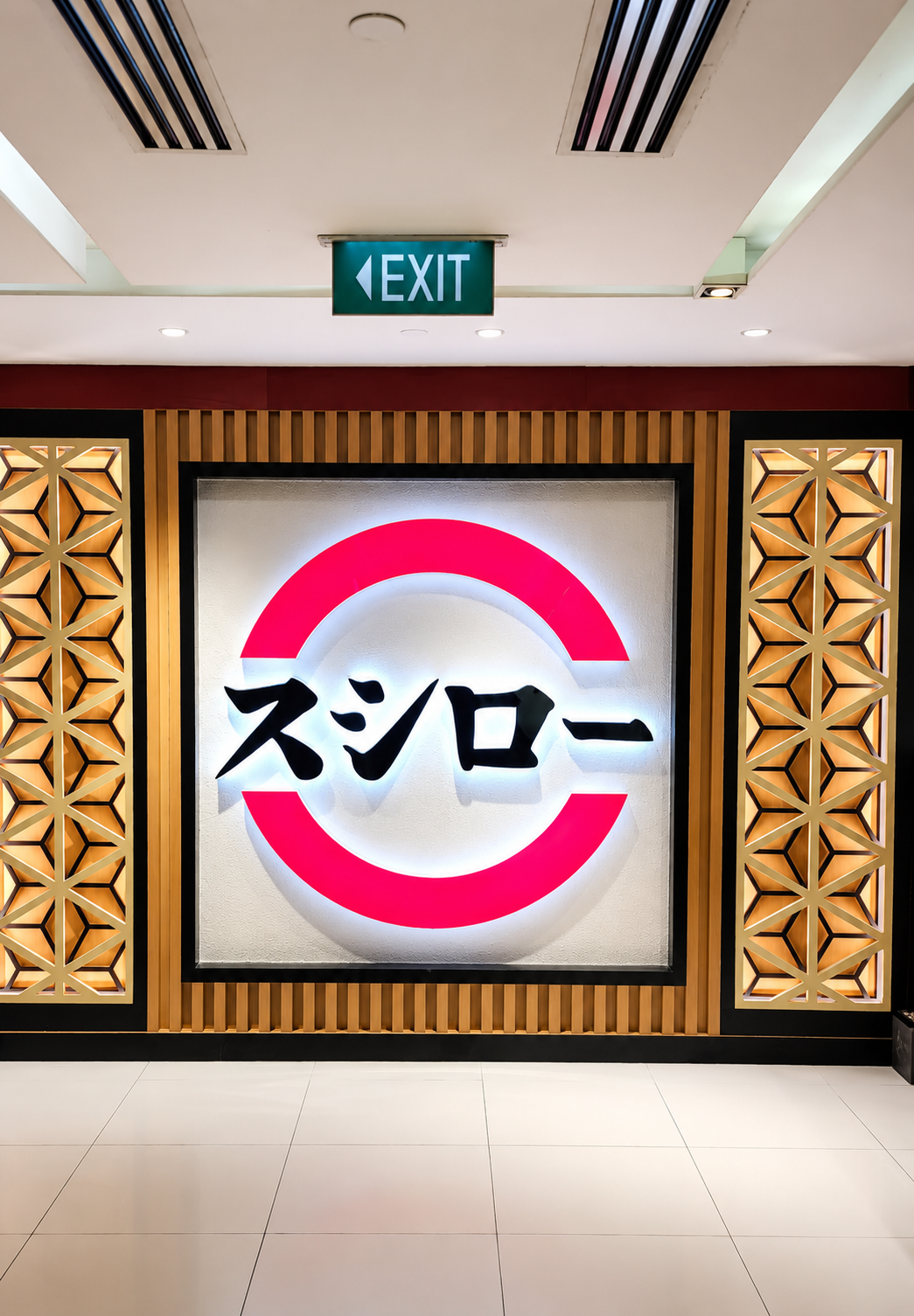 Neon sign with Japanese characters and a red circle design inside a wooden frame, located beneath an illuminated exit sign in an indoor setting.