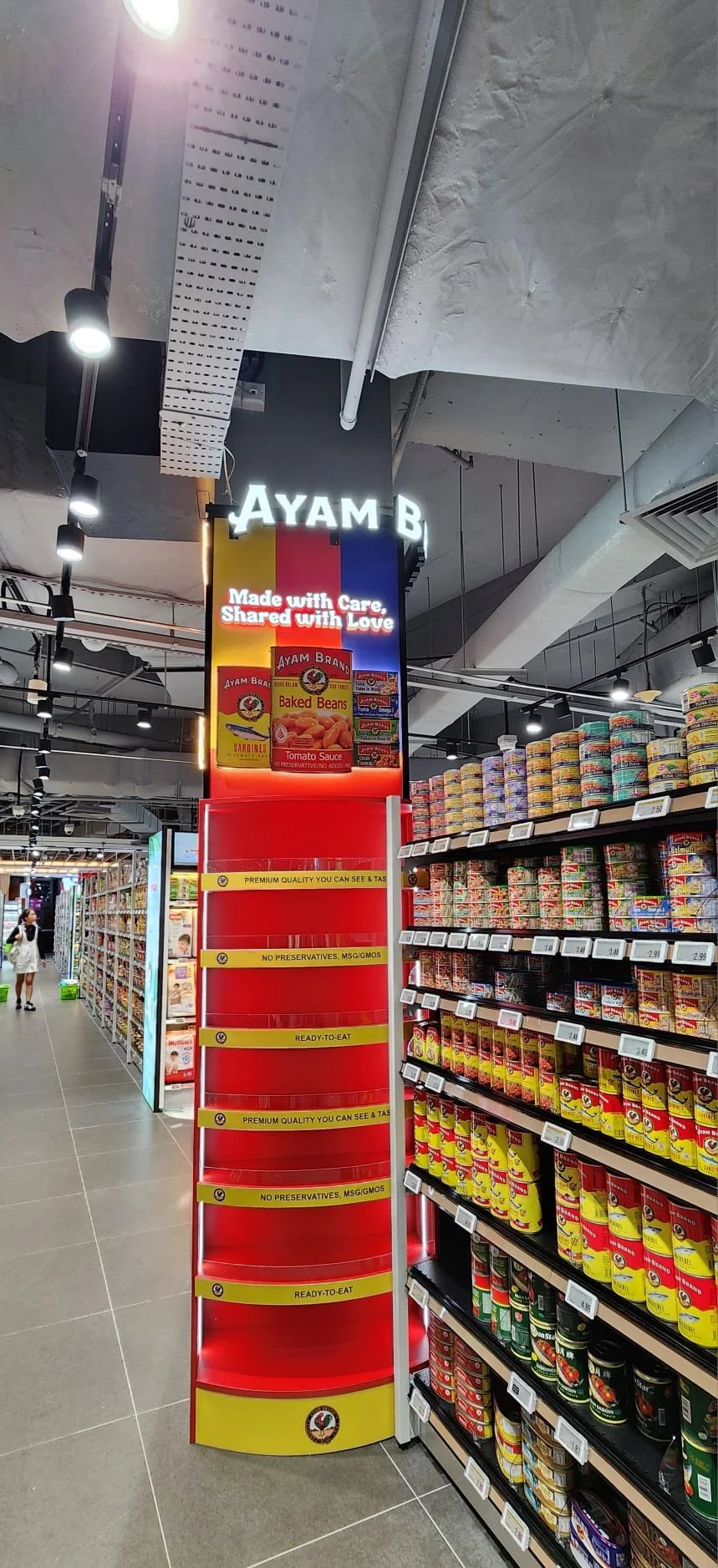 Supermarket aisle with shelves of canned foods, promotional stand for Ayam Brand featuring baked beans and tomato sauce, and a digital sign saying 'Made with Care, Shared with Love'.