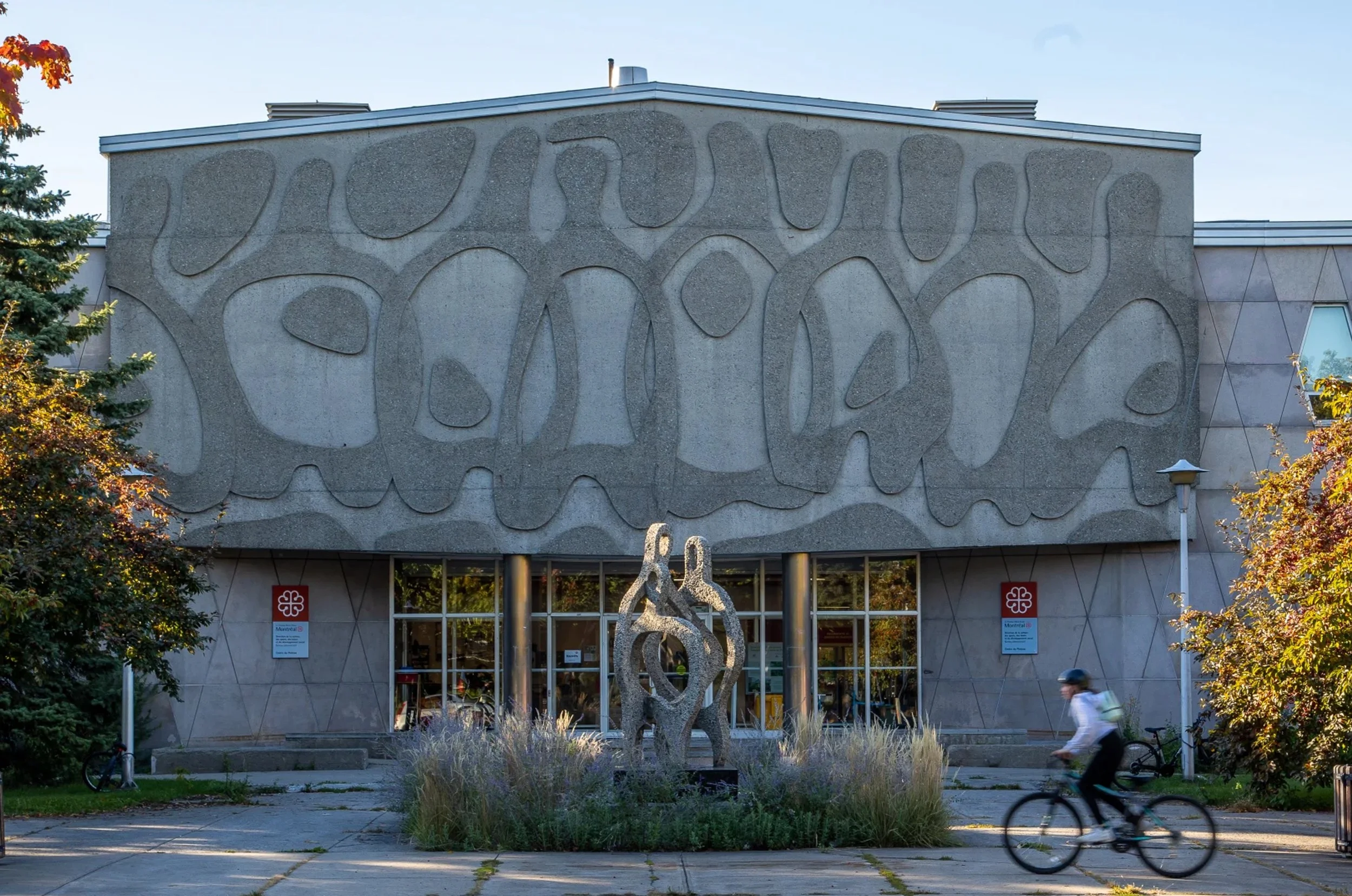 Bâtiment avec une façade en béton décorative, sculptures modernes en métal devant l'entrée, arbres et un cycliste passant devant.