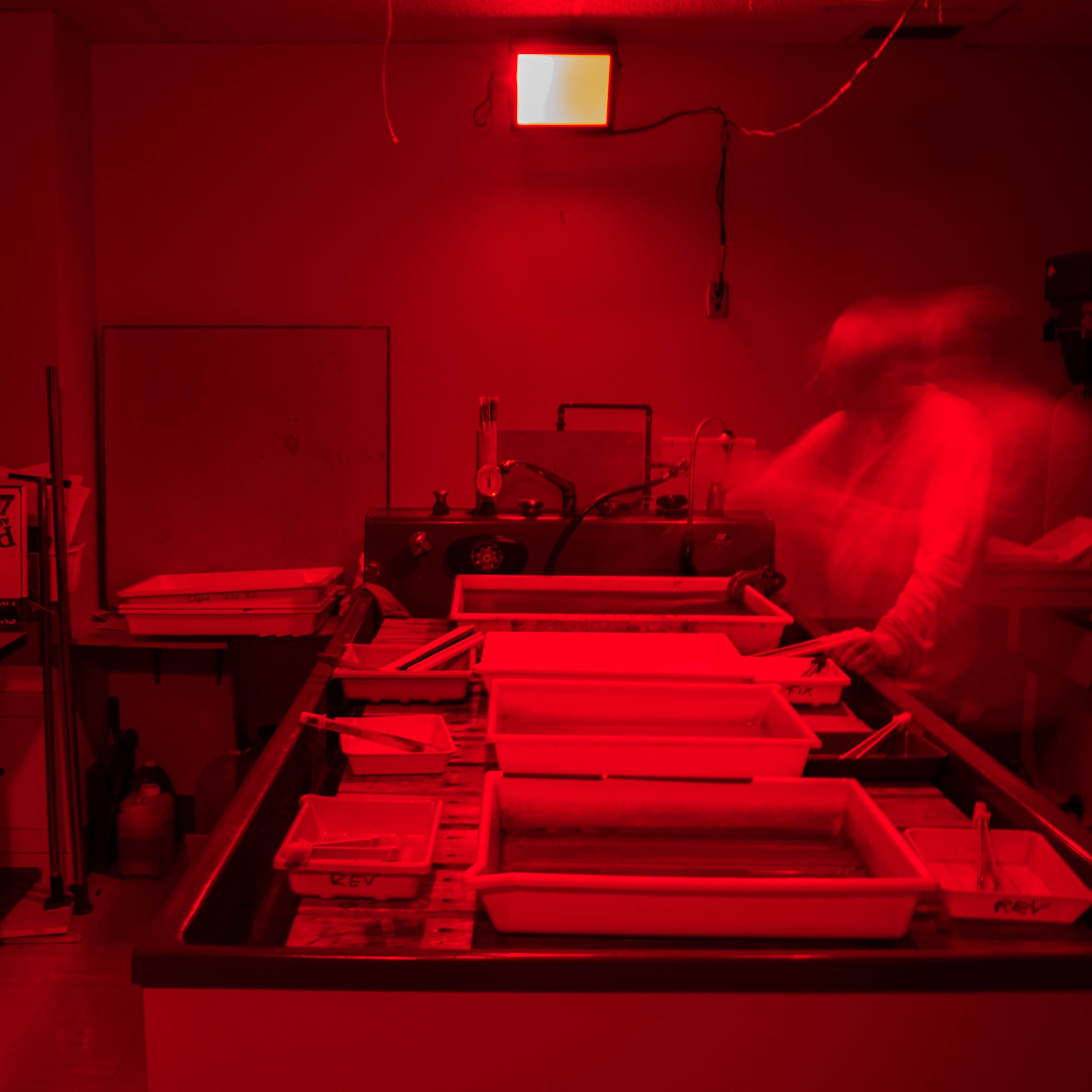 Salle avec éclairage rouge, équipements de laboratoire, table avec boîtes de rangement, personne portant une blouse de laboratoire, lumière rouge au plafond.
