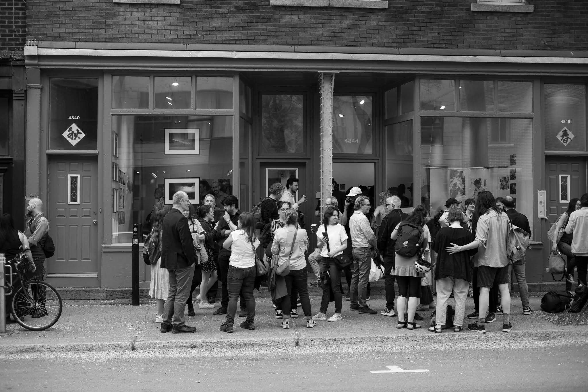 Une foule de personnes se tient devant une devanture de magasin ou d'exposition, visible à travers une grande vitrine. La scène est en noir et blanc.