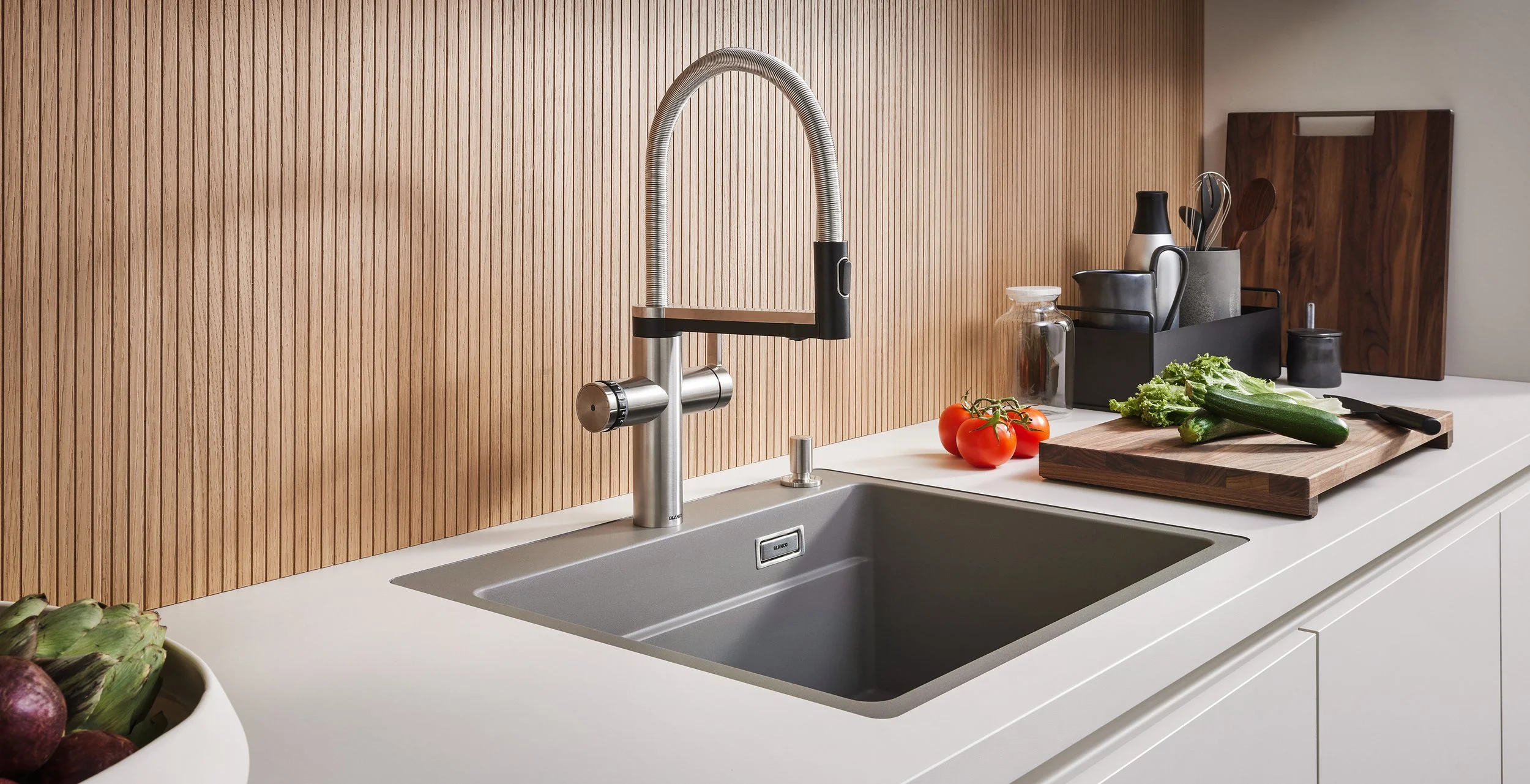 Modern kitchen with a white countertop, stainless steel sink, and a wooden backsplash. There are fresh vegetables including tomatoes, zucchini, and lettuce, along with kitchen utensils and a chopping board.