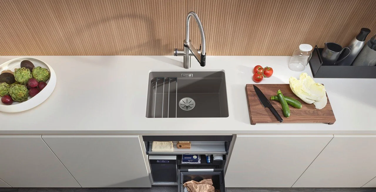 Kitchen countertop with a bowl of artichokes and radishes on the left, a stainless steel sink with a faucet in the center, and a cutting board with cucumbers, tomatoes, and cabbage on the right.