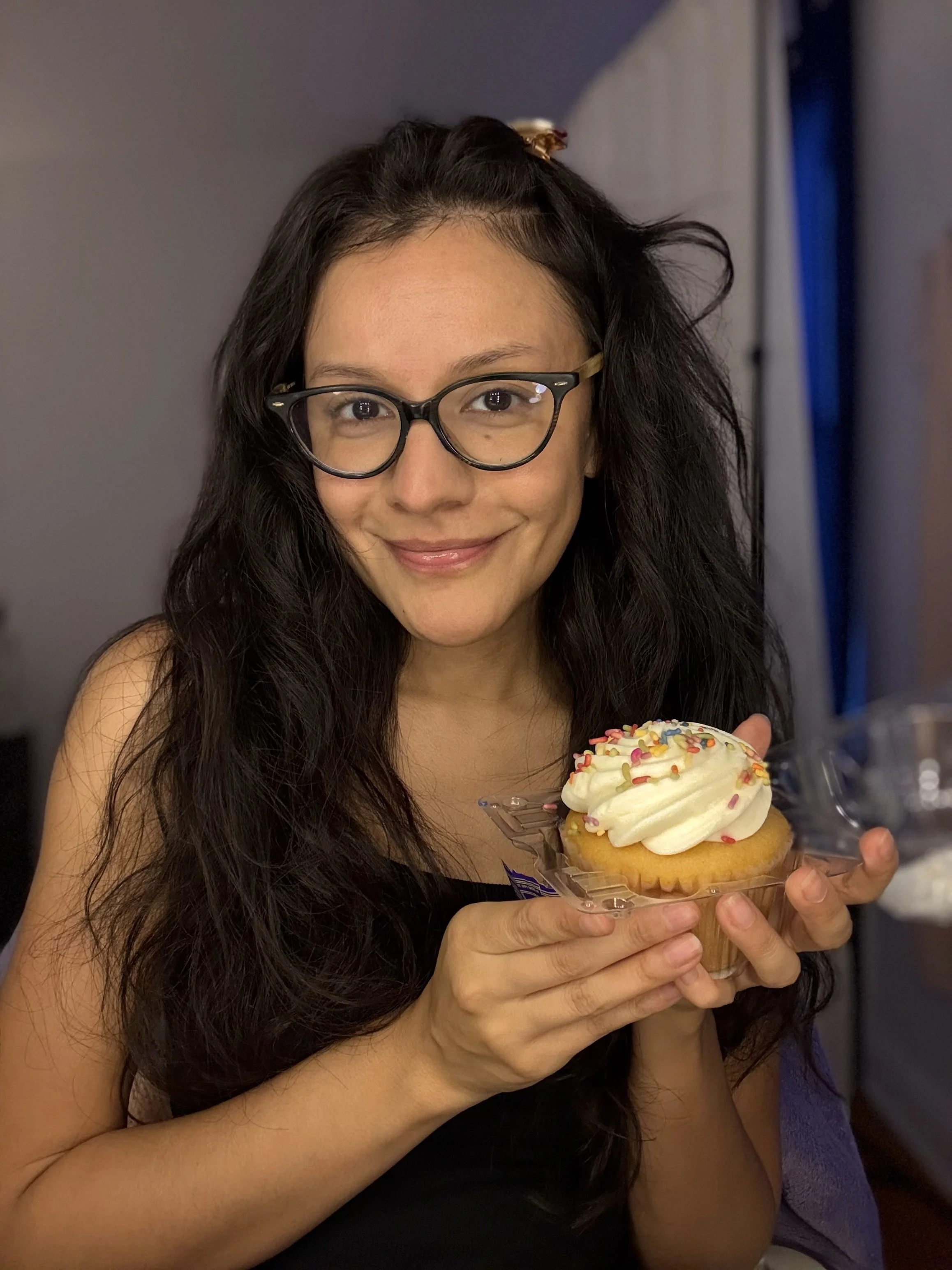 A woman with long dark hair and glasses holding a cupcake with white frosting and colorful sprinkles.