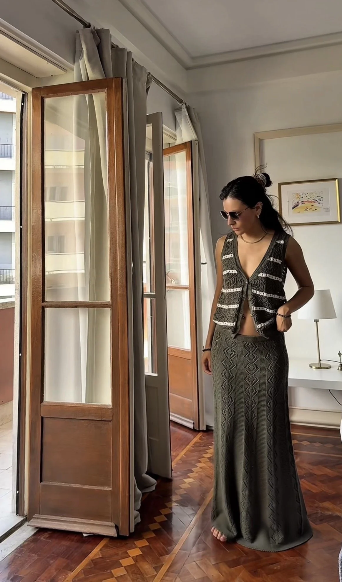 A woman with long dark hair, wearing sunglasses, a sleeveless patterned vest, and wide-leg patterned pants, stands indoors near open glass doors with curtains, looking down. There is framed artwork on the wall, a white lamp on a bedside table, and a wooden floor with a decorative pattern.