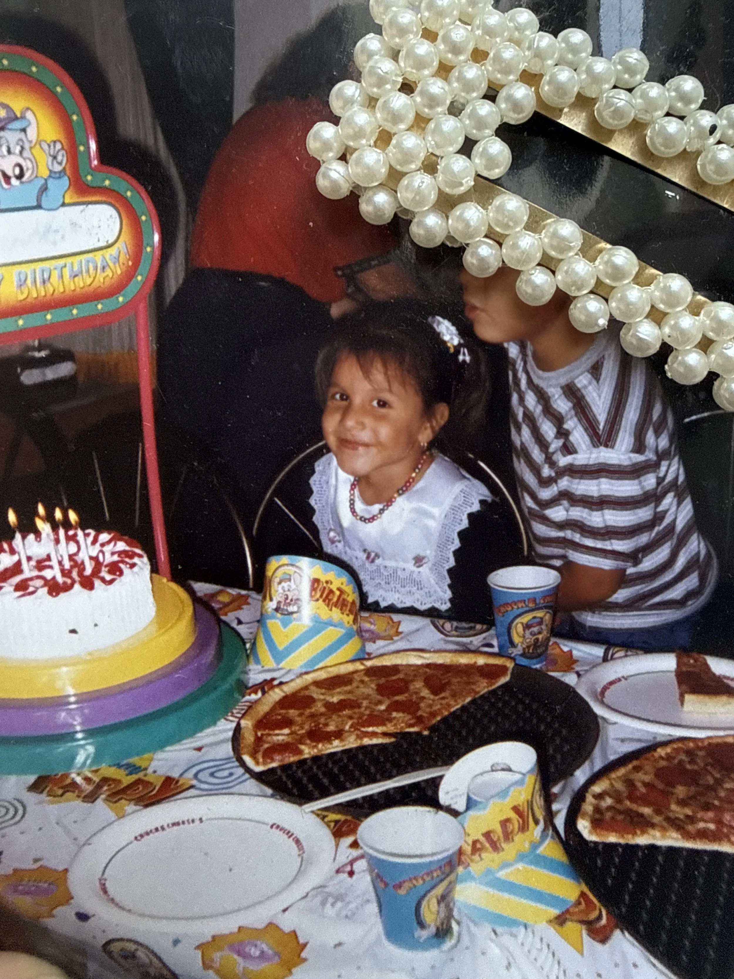 A birthday celebration with a cake, pizza, and party decorations. Two children are present, one smiling at the camera and the other leaning over. There are birthday hats, themed paper cups, and pearl necklaces on the table.
