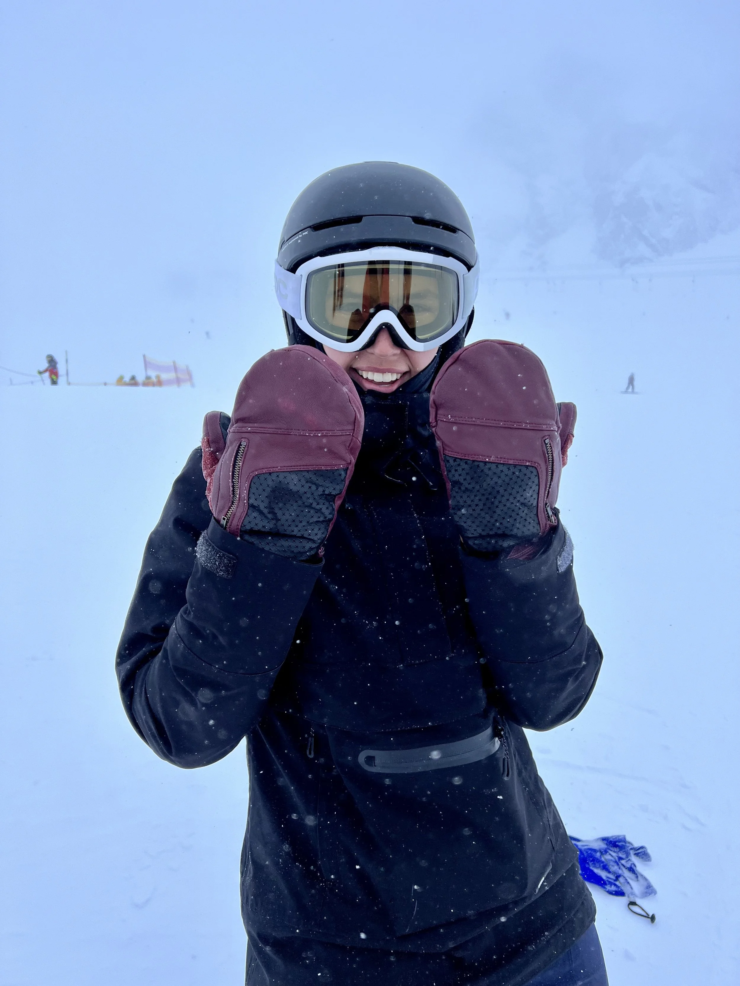 A person in winter clothing, wearing a helmet and goggles, is smiling and showing off Swany Mittens in the cold. 