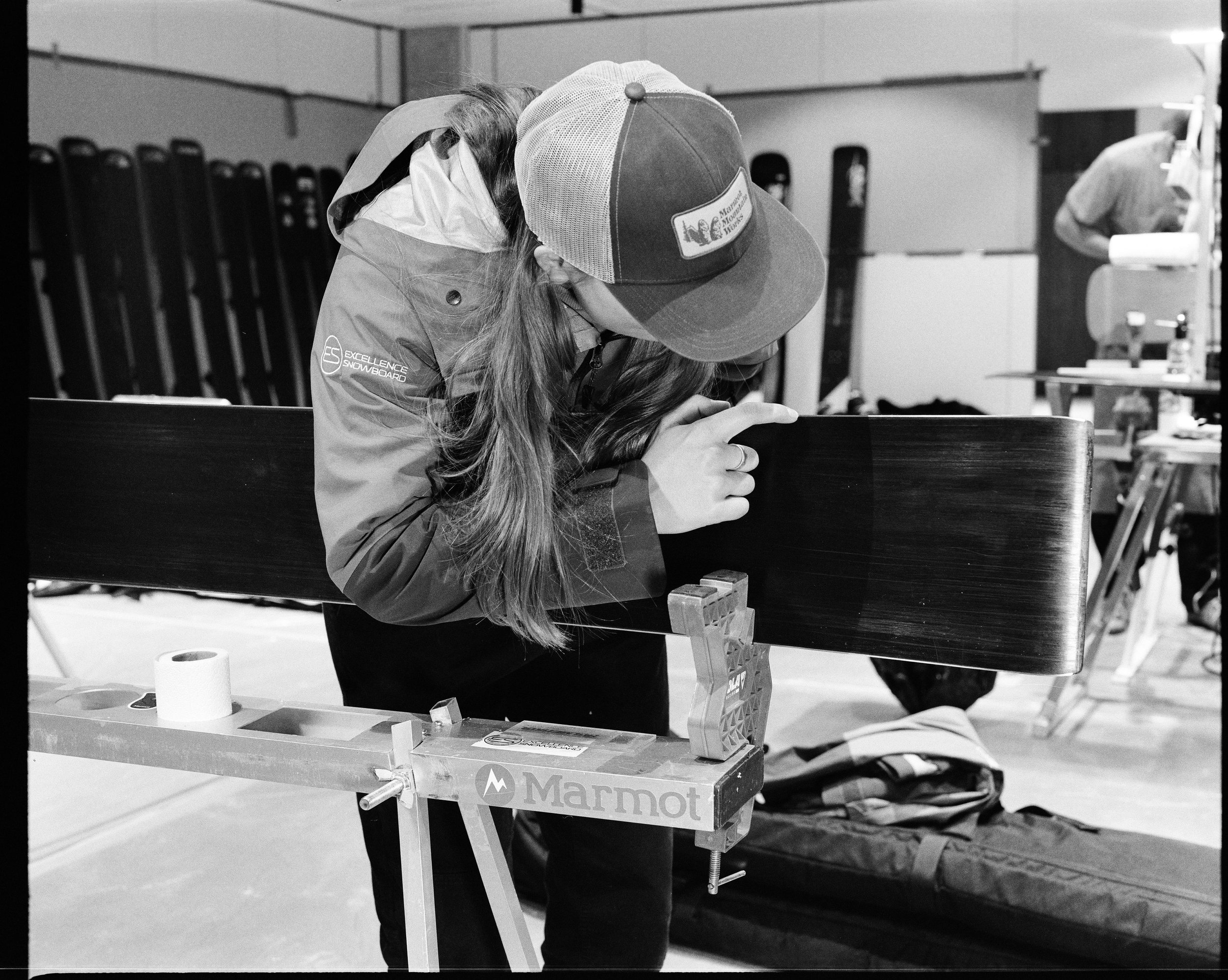 Person leaning over a table, wearing a cap and jacket, working on snowboard preparation at Marmot branded workbench in an indoor setting.
