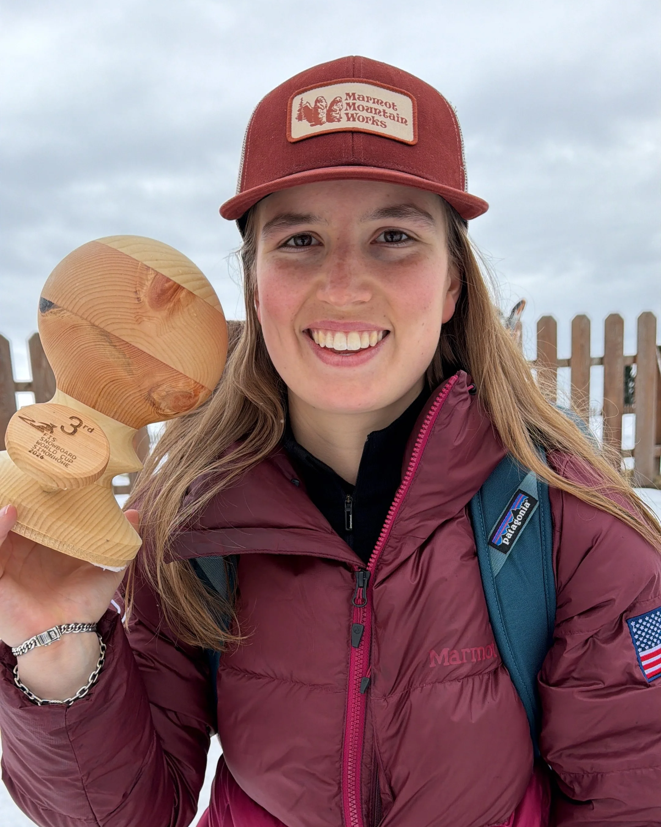 A woman smiling while holding a wooden trophy with a winter sports award engraving. She is outdoors, wearing a red Marmot jacket and a red hat with a Marmot Mountain Works logo.